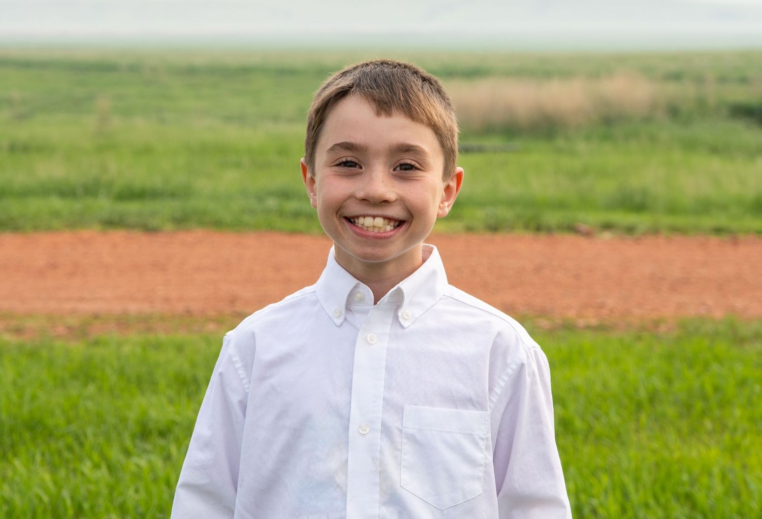Smiling boy in white button-up shirt posing in a green field.