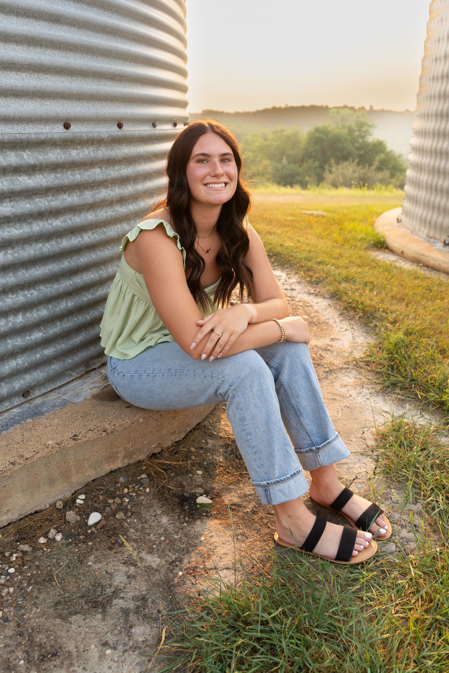 Smiling young woman in green top and jeans sits beside corrugated metal grain bins at sunset.