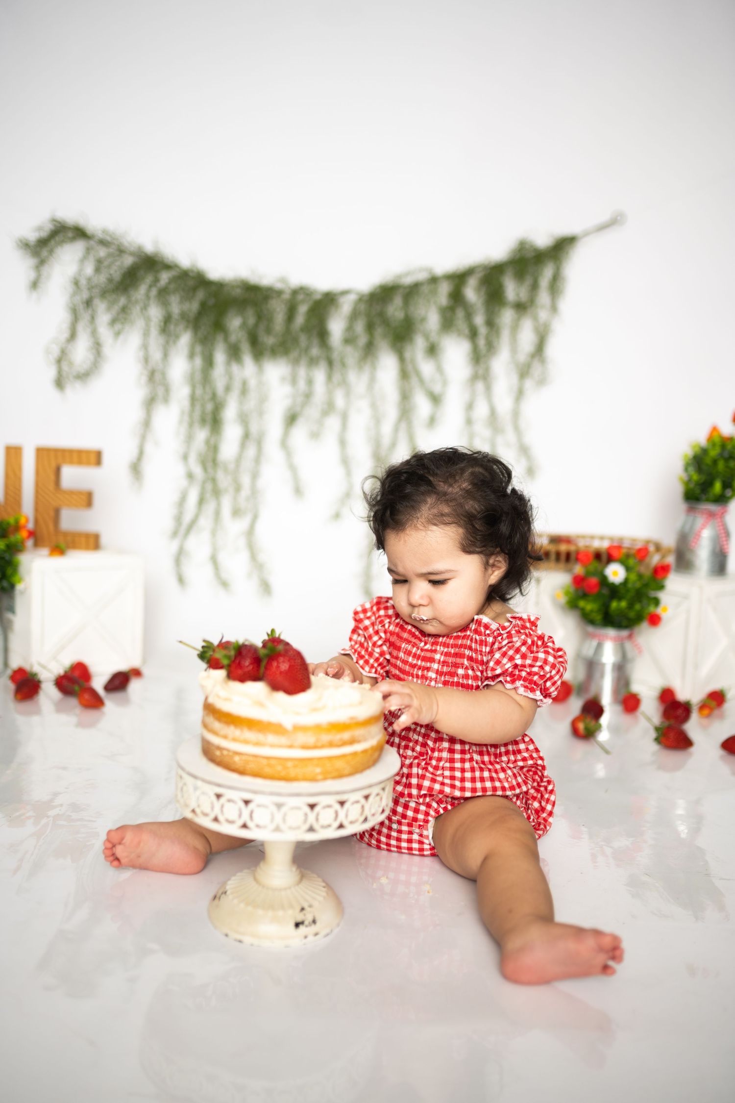 Baby in red gingham dress reaching for cake on white pedestal with strawberry decorations.