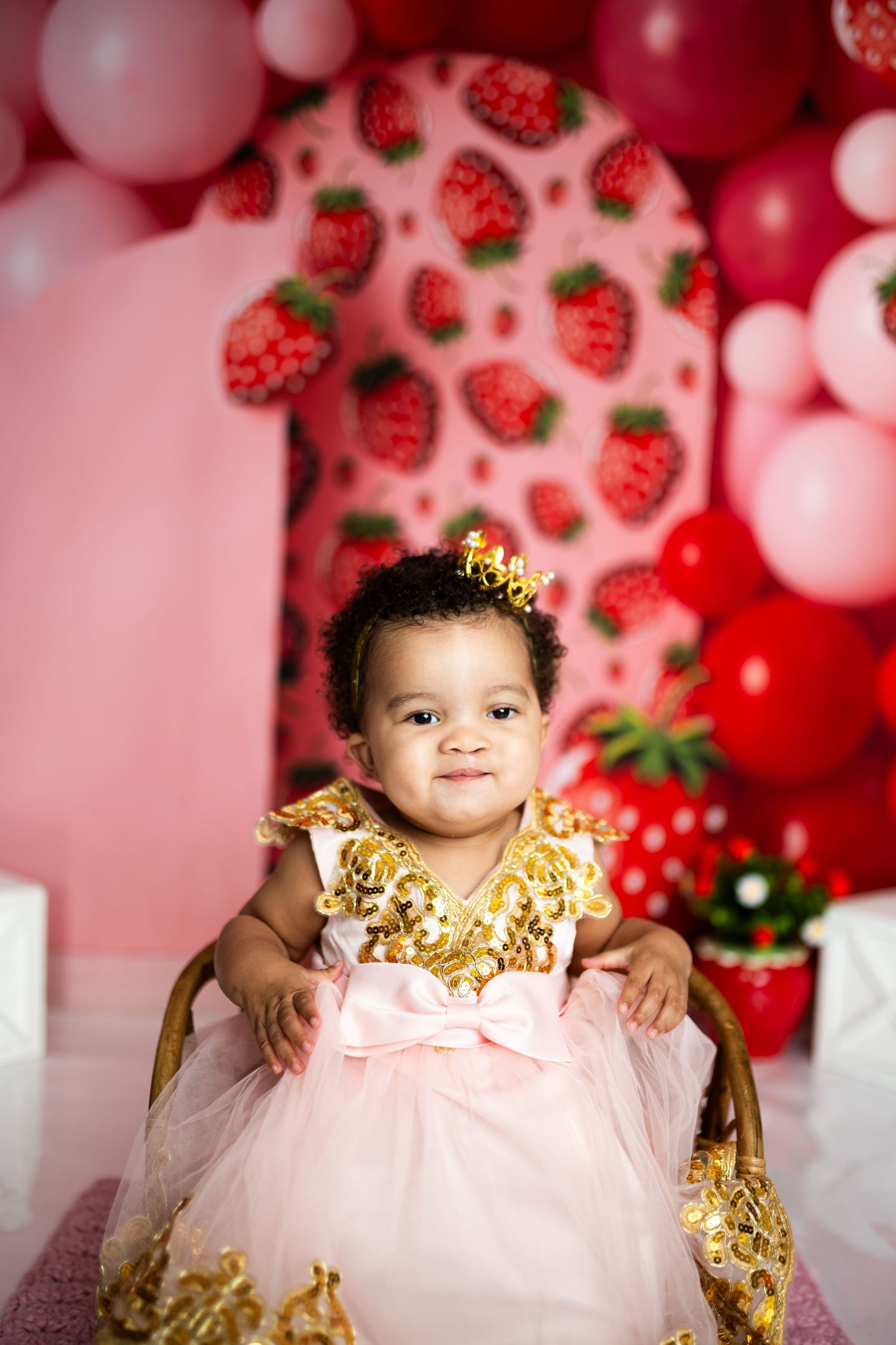 Baby in pink and gold dress against strawberry balloon backdrop for first birthday photos.