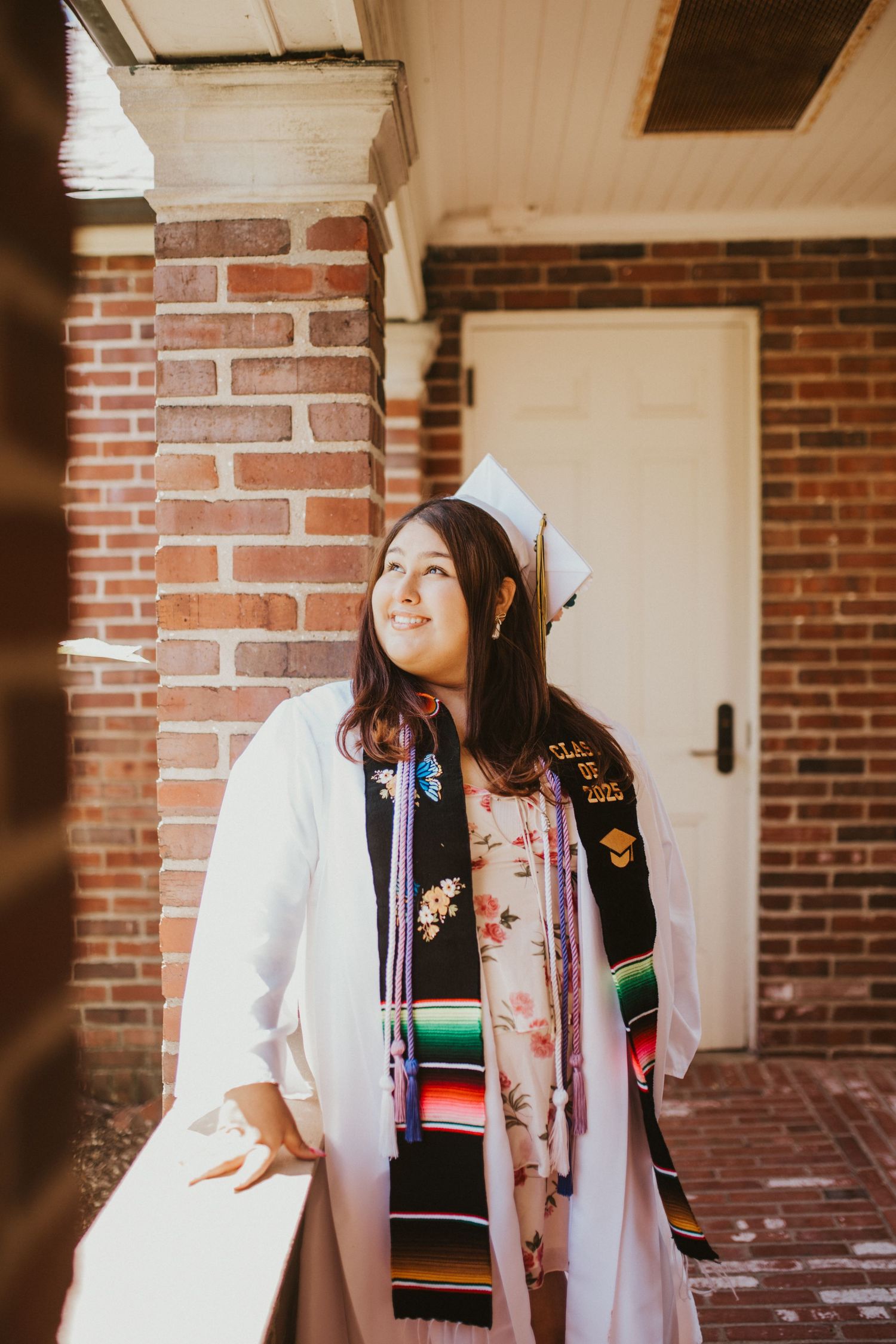 Graduate in white coat and colorful stole poses against brick building.
