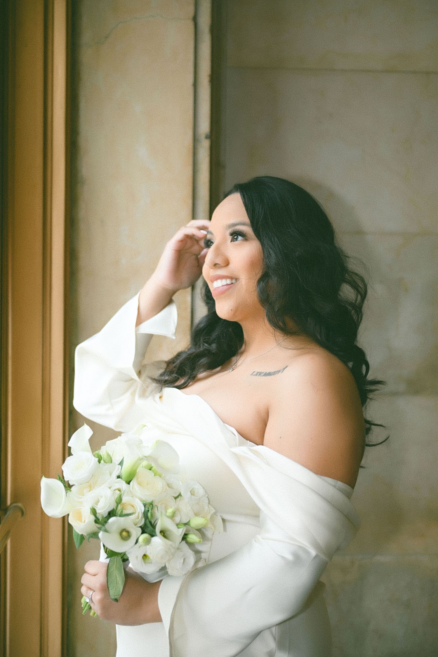 A bride in a white dress holds a bouquet of white flowers while posing near a window.