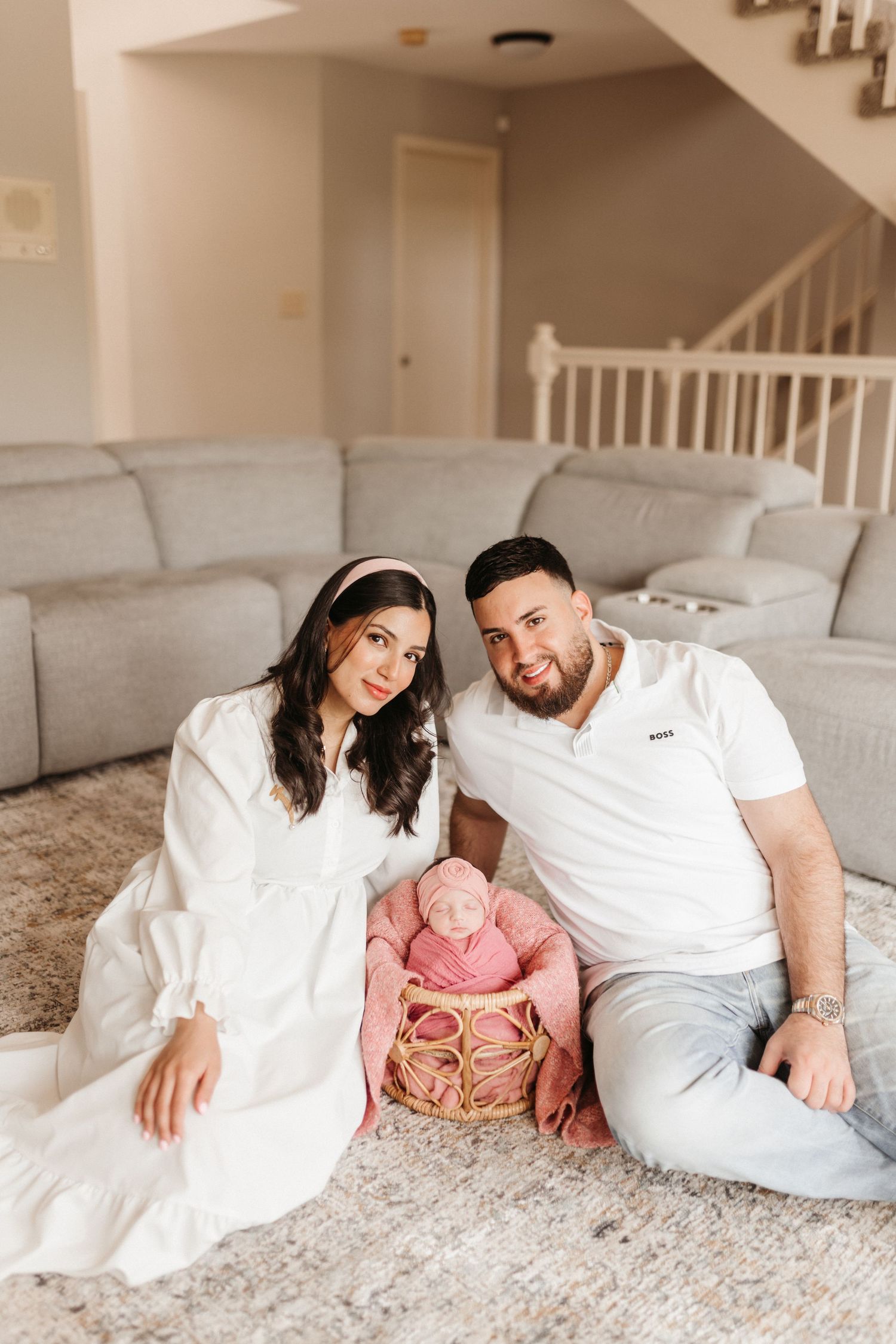 A couple sits on a carpet with their newborn baby wrapped in pink in a wicker basket in a cozy living room.