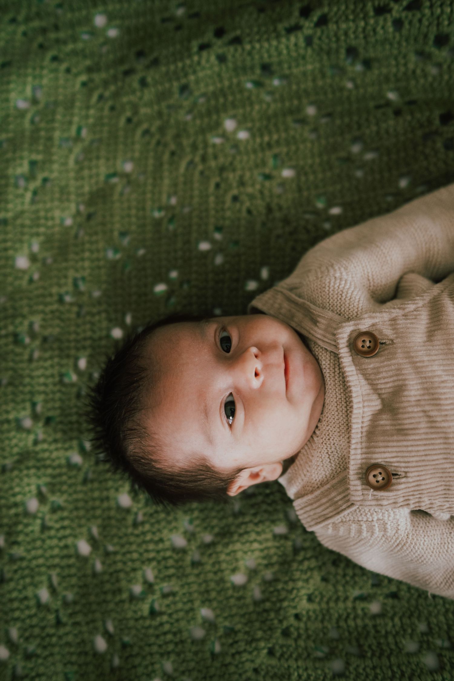 Newborn baby lying on a green knit blanket wearing a beige button cardigan, looking up with bright eyes.