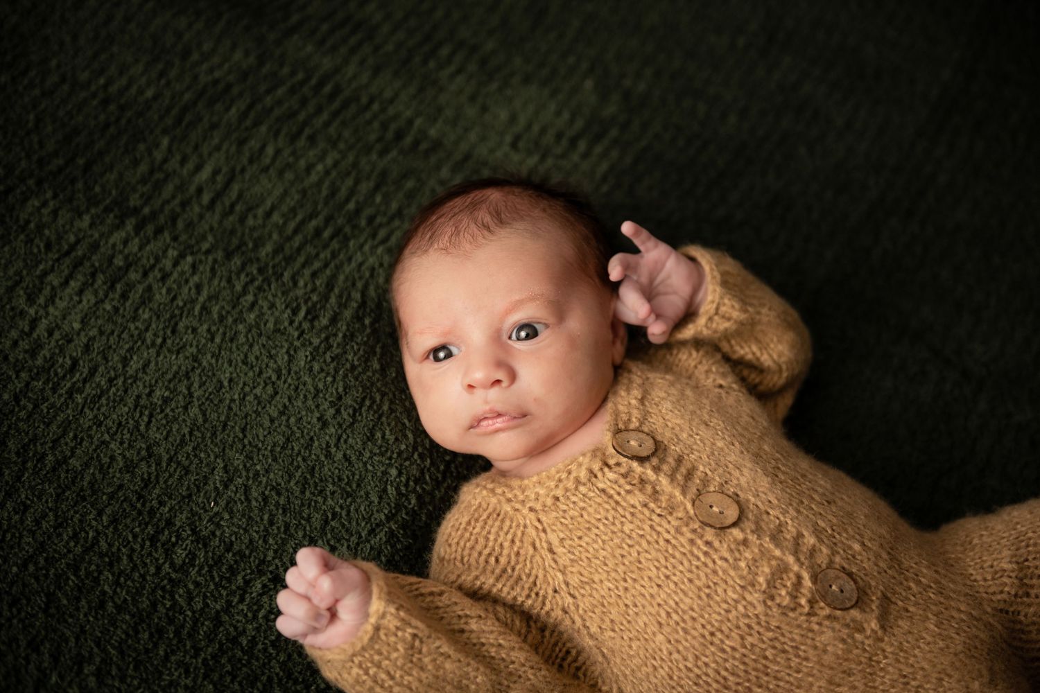 Newborn baby in golden knit romper lying on green blanket, looking up with bright blue eyes.