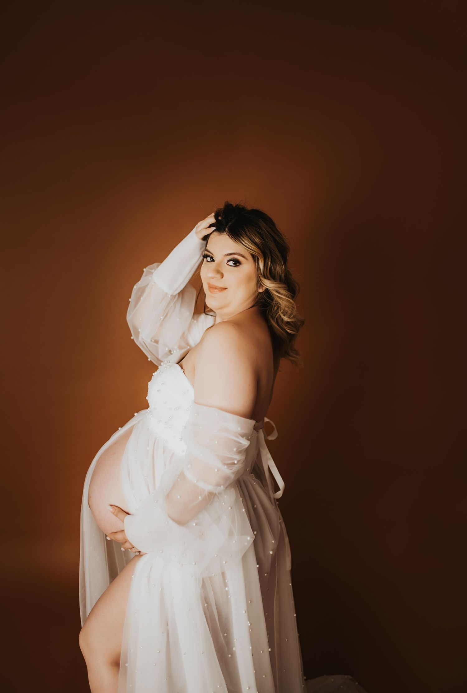 Maternity portrait of woman in flowing white dress against warm brown background.