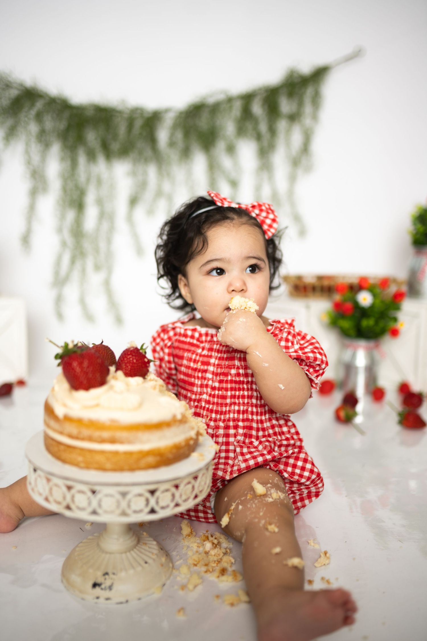 Baby enjoying cake smash session with white cake stand and strawberry themed decorations.