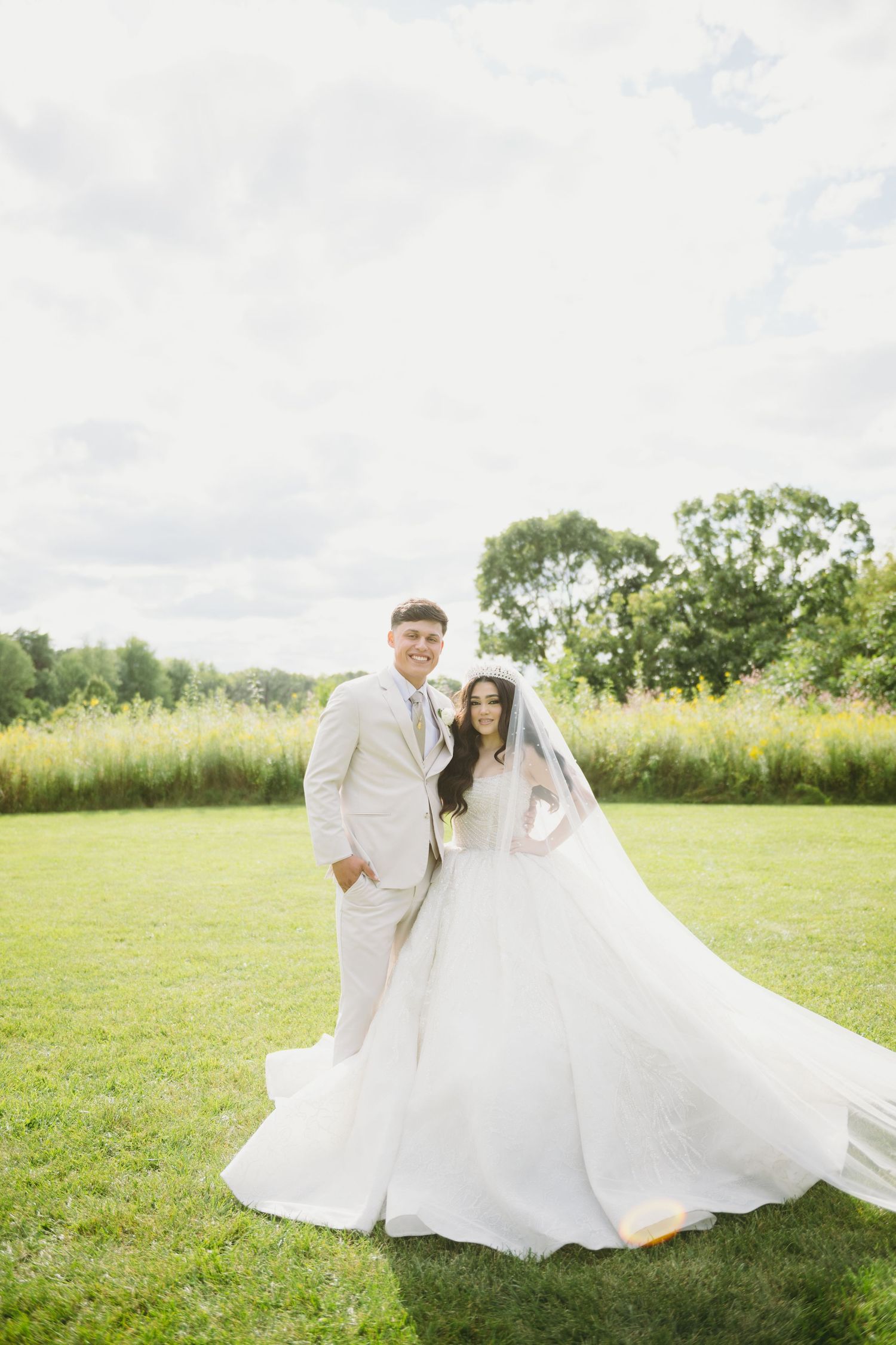 Newlyweds in white attire twirling in a sunny meadow.