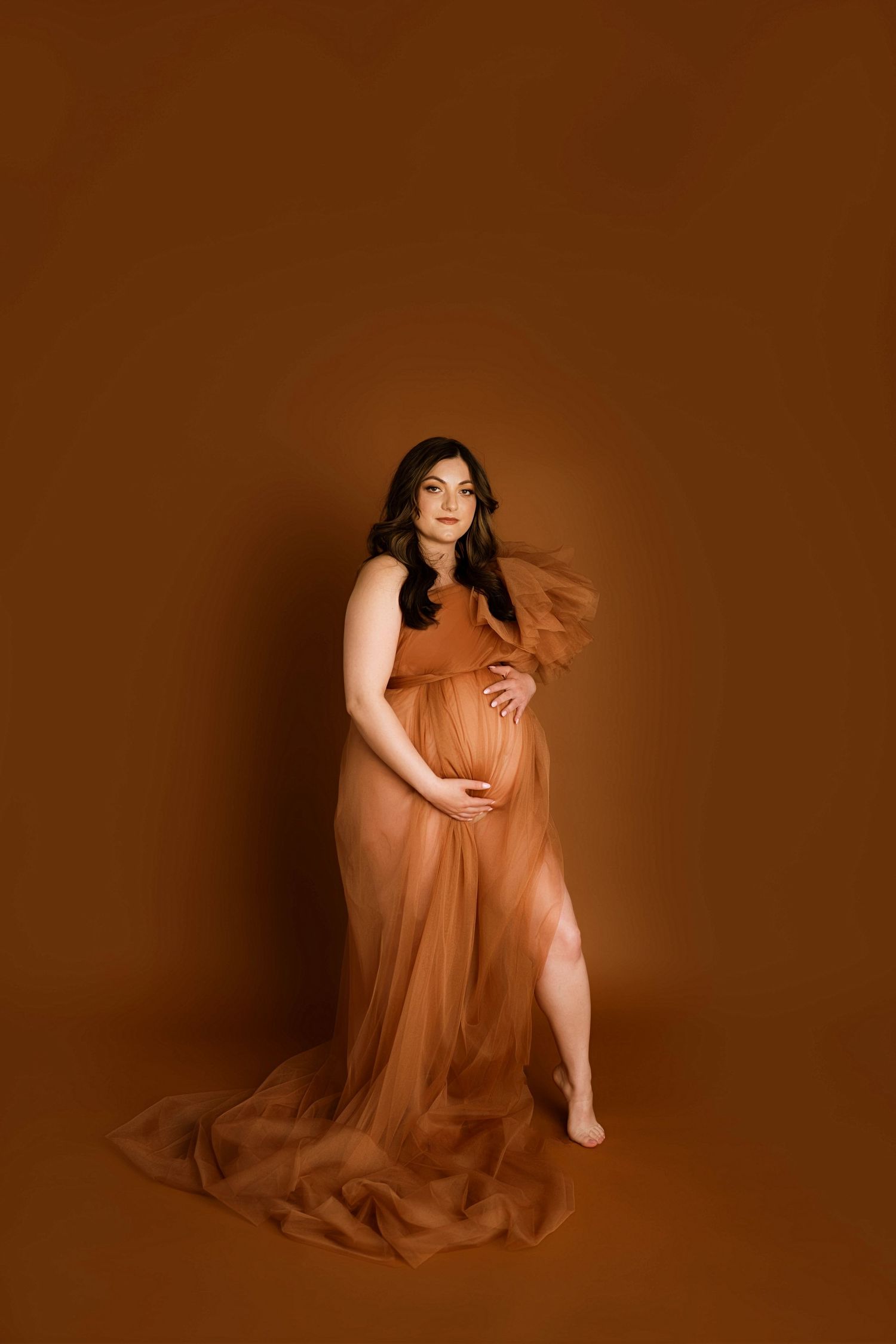 Pregnant woman in flowing rust-colored tulle gown poses gracefully against warm brown studio backdrop.