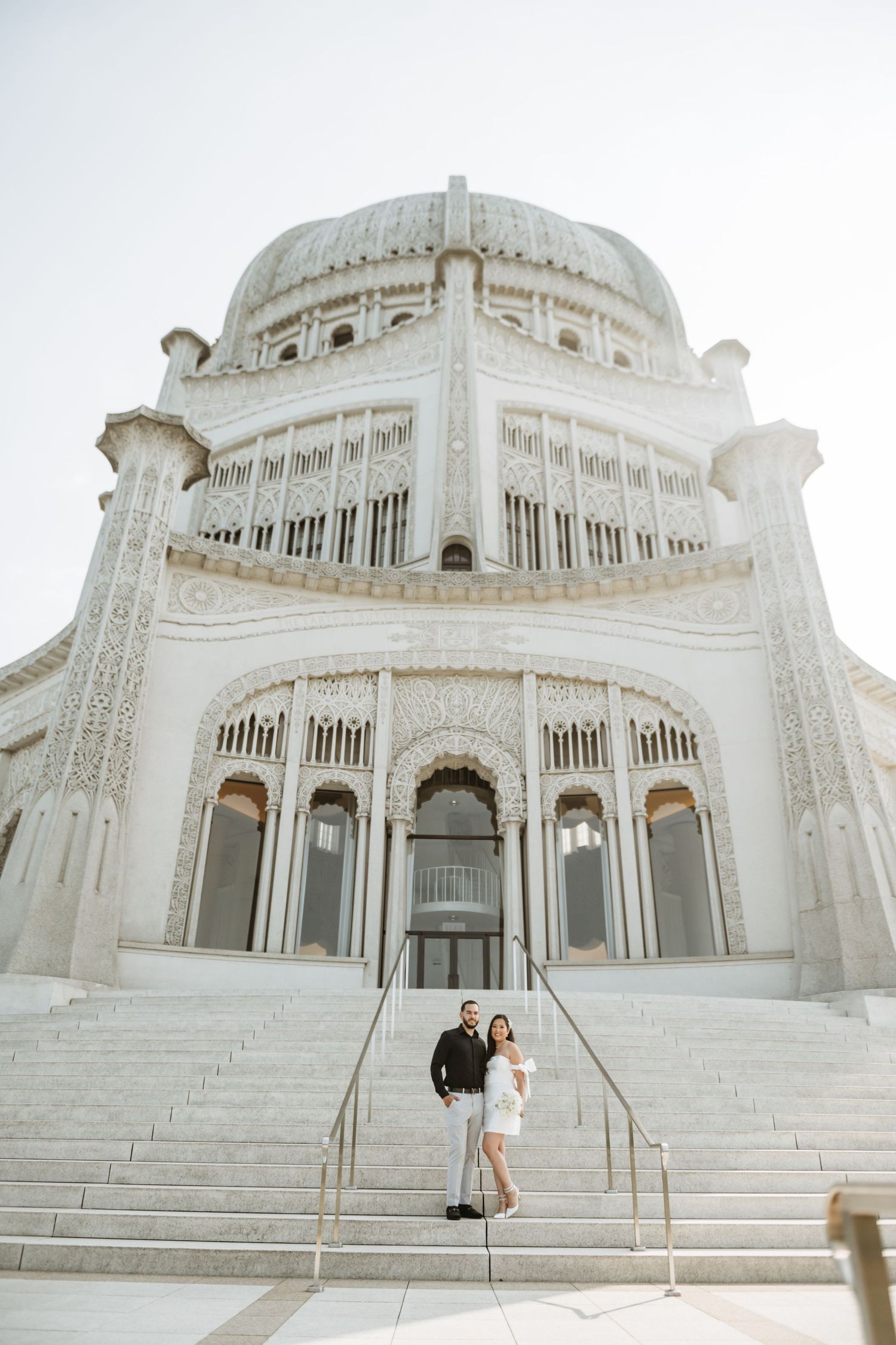 A couple poses on the grand steps of a stunning white domed building with ornate architecture.
