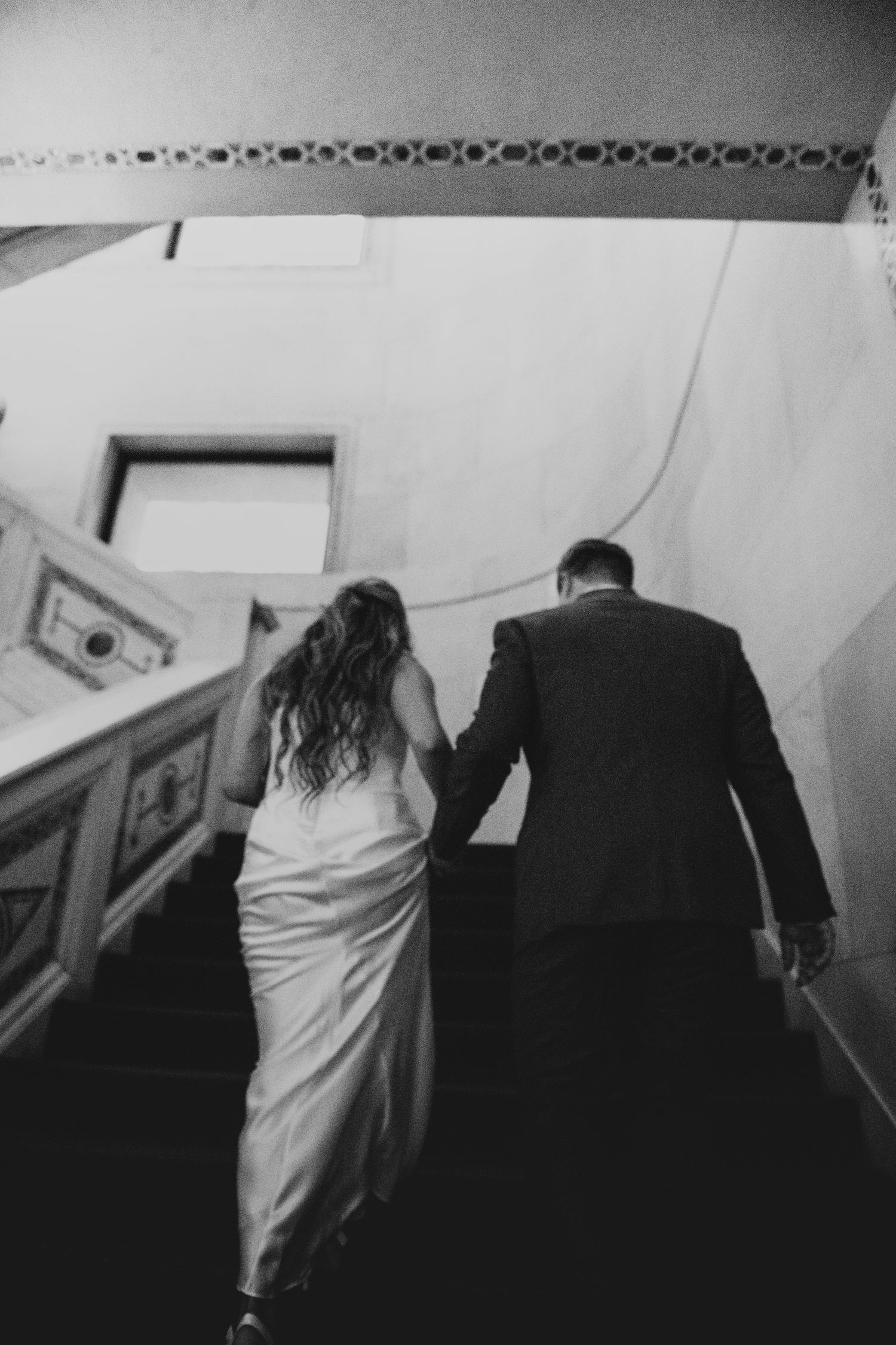 Black and white photograph of a couple walking up stairs in formal wedding attire.
