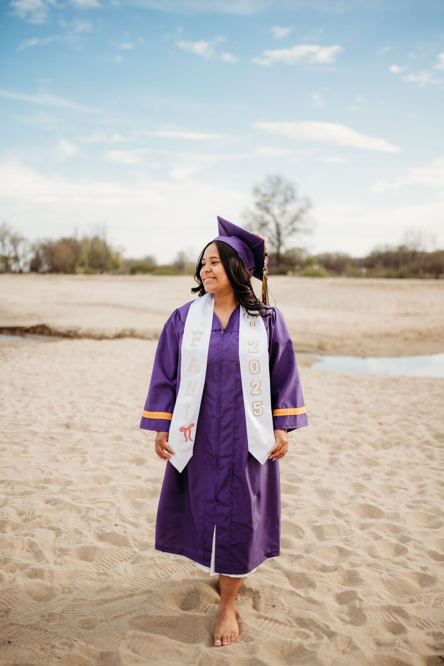 Graduate in purple cap and gown sits thoughtfully on sandy beach during outdoor graduation photoshoot.