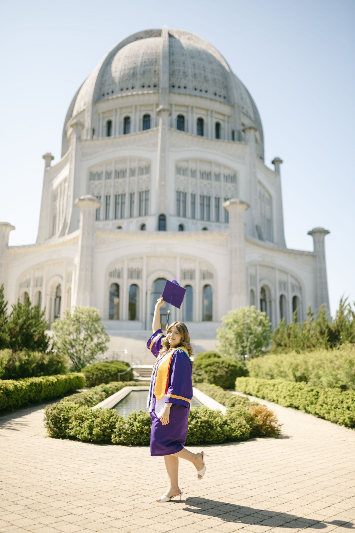 Graduate in purple cap and gown jumps joyfully in front of a grand domed building with manicured gardens.