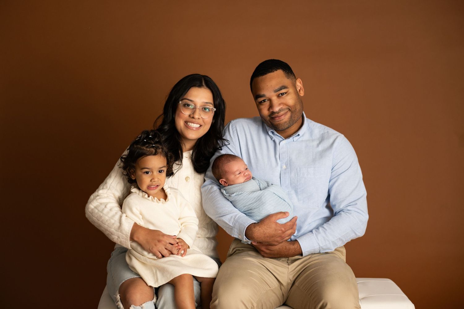 A family of four poses for a newborn portrait, parents holding a toddler and swaddled baby against a warm brown backdrop.