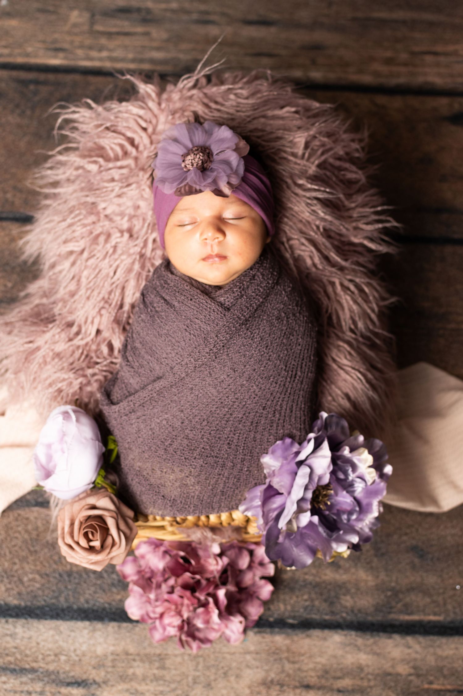 Newborn baby girl swaddled in purple wrap with floral headband, sleeping peacefully in fluffy lavender basket with flowers.