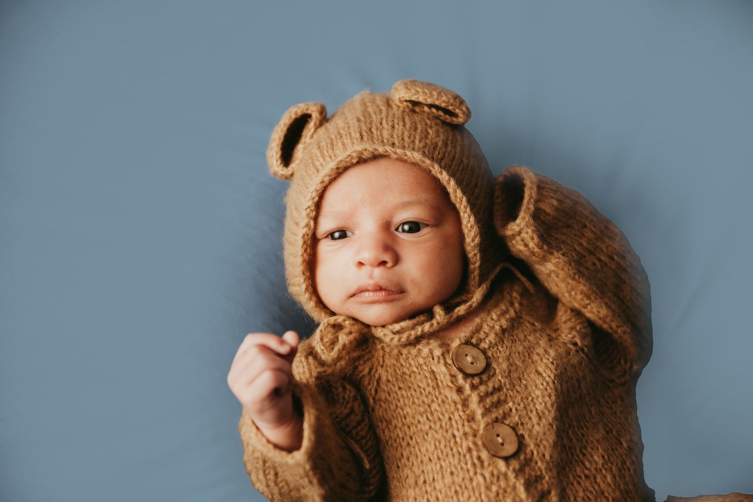 Newborn baby wearing a cozy brown knit bear onesie and bonnet with ears, lying on a soft blue background.