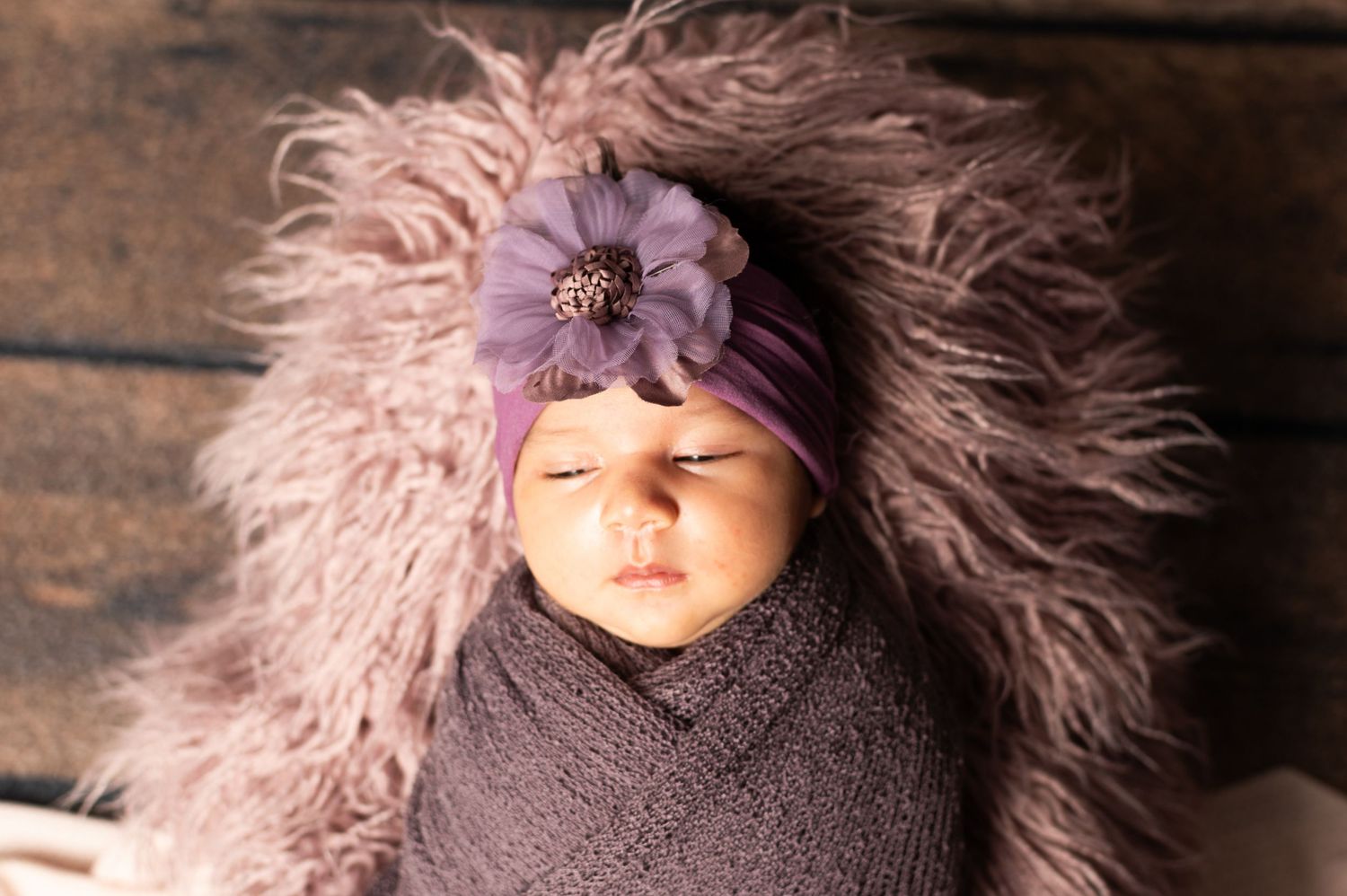 Sleeping newborn baby swaddled in purple wrap with floral headband on fluffy lavender fur backdrop.