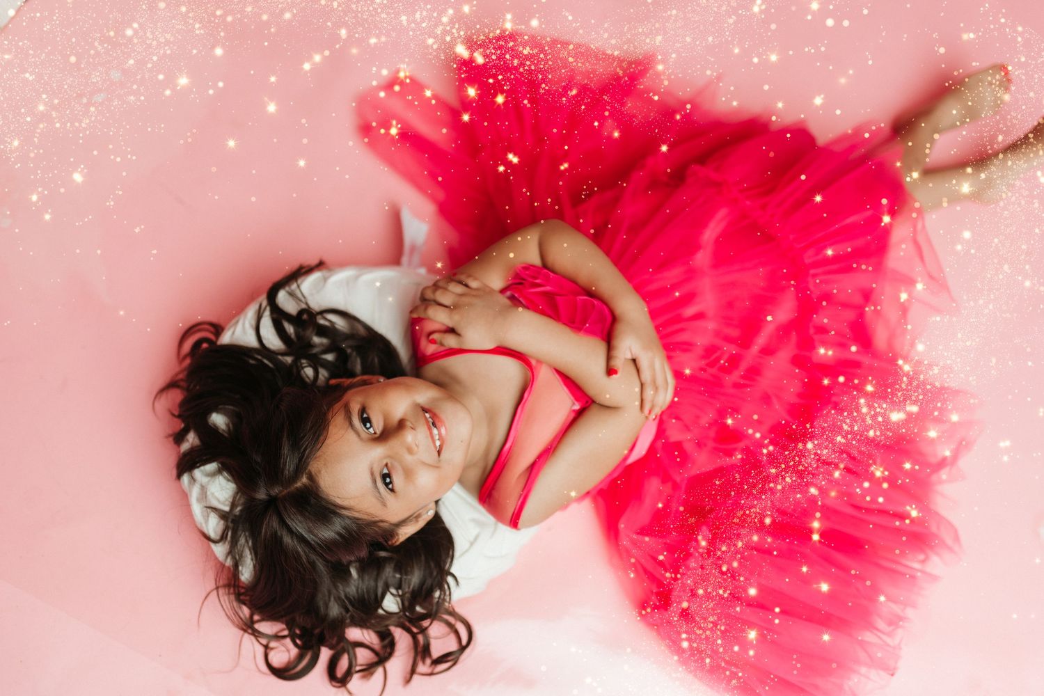 Person in a flowing red tulle dress lying against pink background with sparkles.
