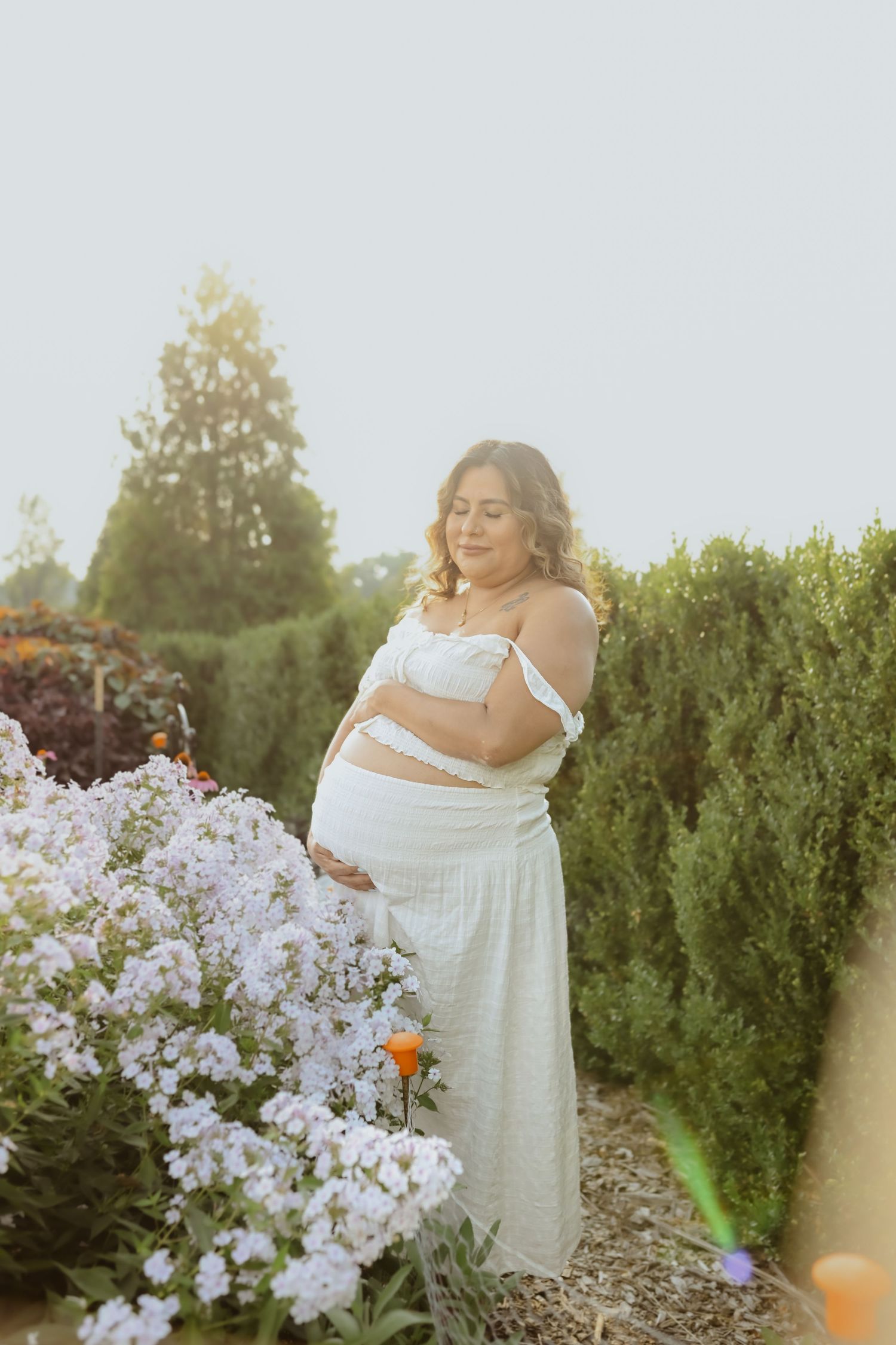 Ethereal outdoor maternity photos with white flowers and sunlit garden setting.