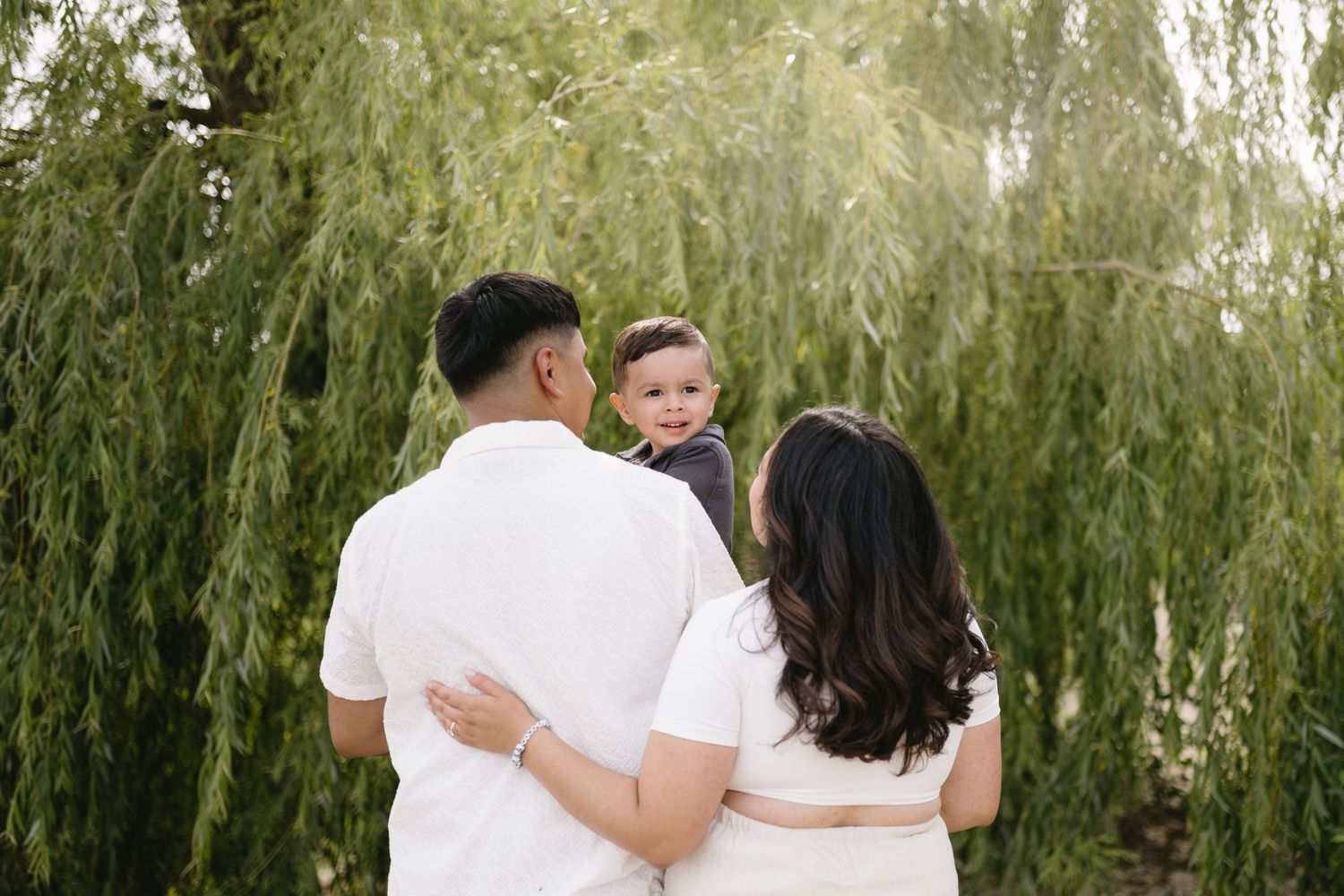 Family in white outfits embracing while standing under a weeping willow tree.
