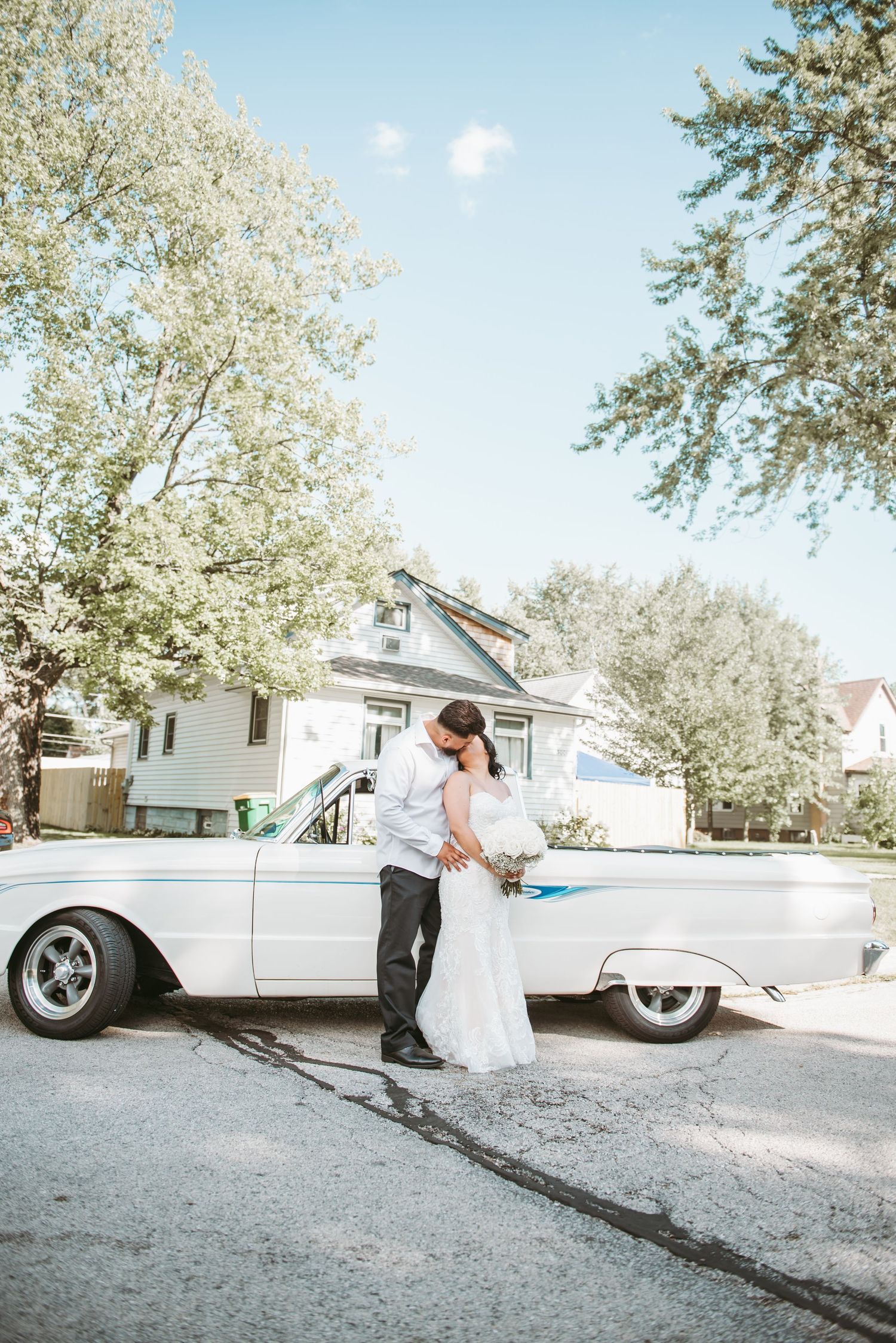 A couple poses romantically next to a classic white vintage car on a sunny day.