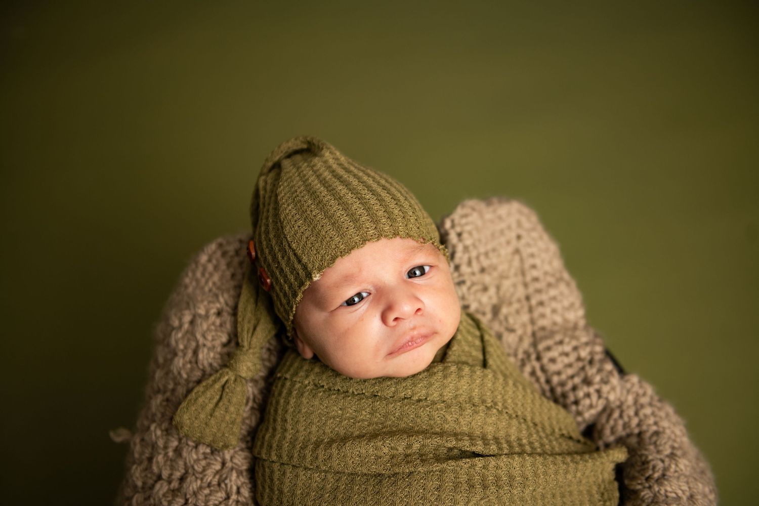 Newborn baby wrapped in green knit blanket and matching hat, posed in a basket against a green background.
