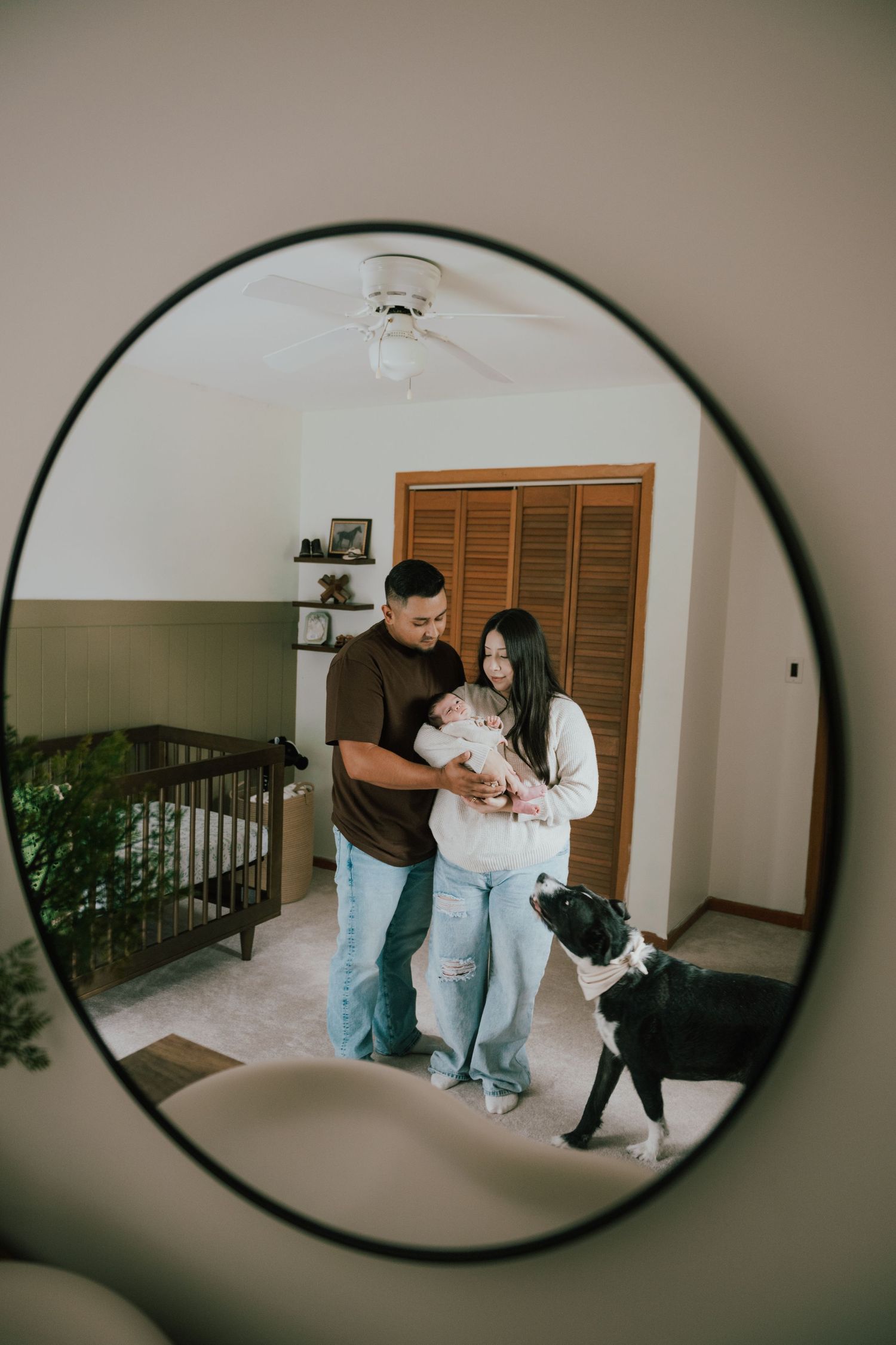 A couple holds their newborn baby in a nursery, reflected in a round mirror, with a black and white dog nearby.