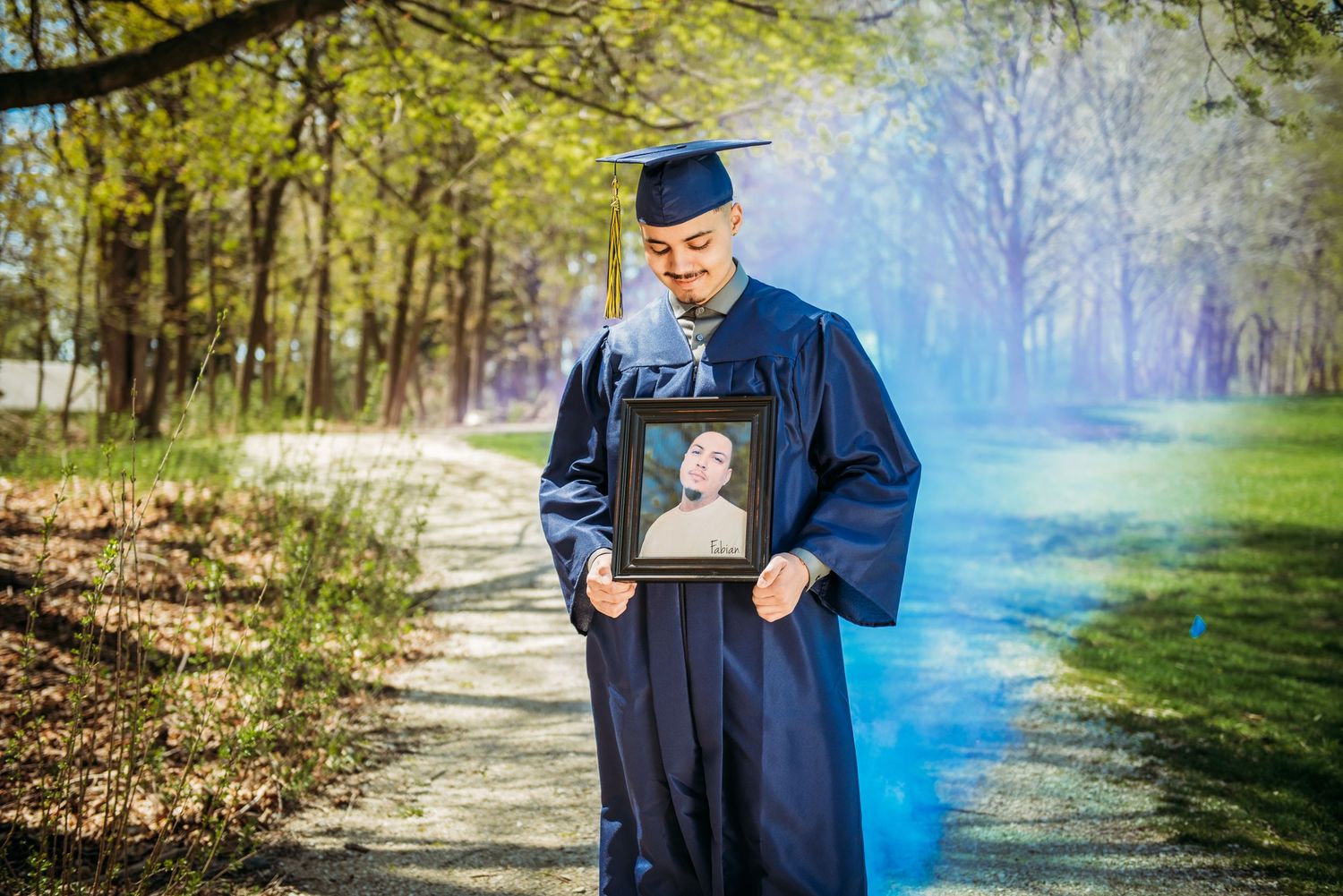 Graduate in blue cap and gown holding blue smoke bomb in outdoor wooded setting.