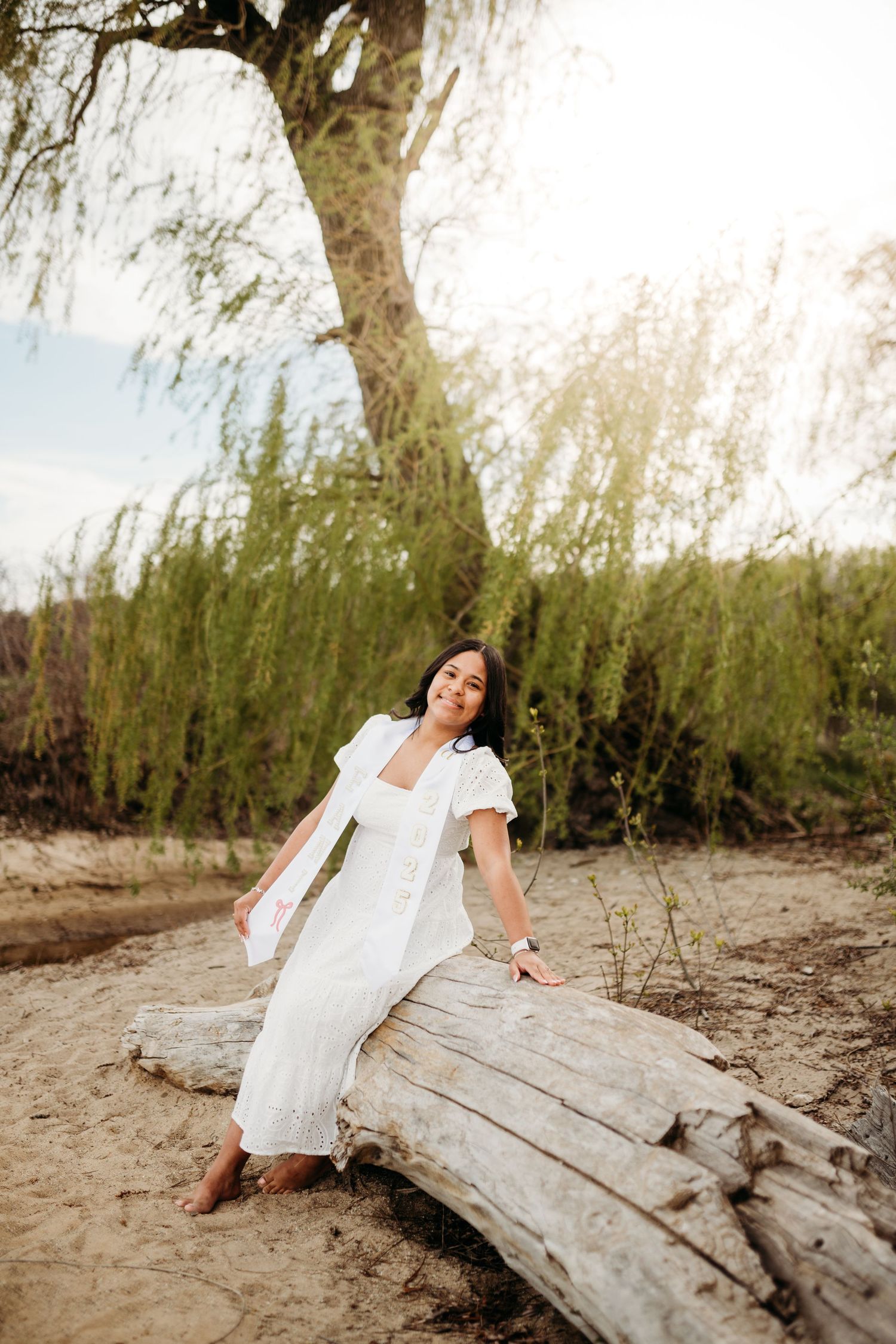 A white dress photoshoot on a sandy beach with driftwood and willow trees.