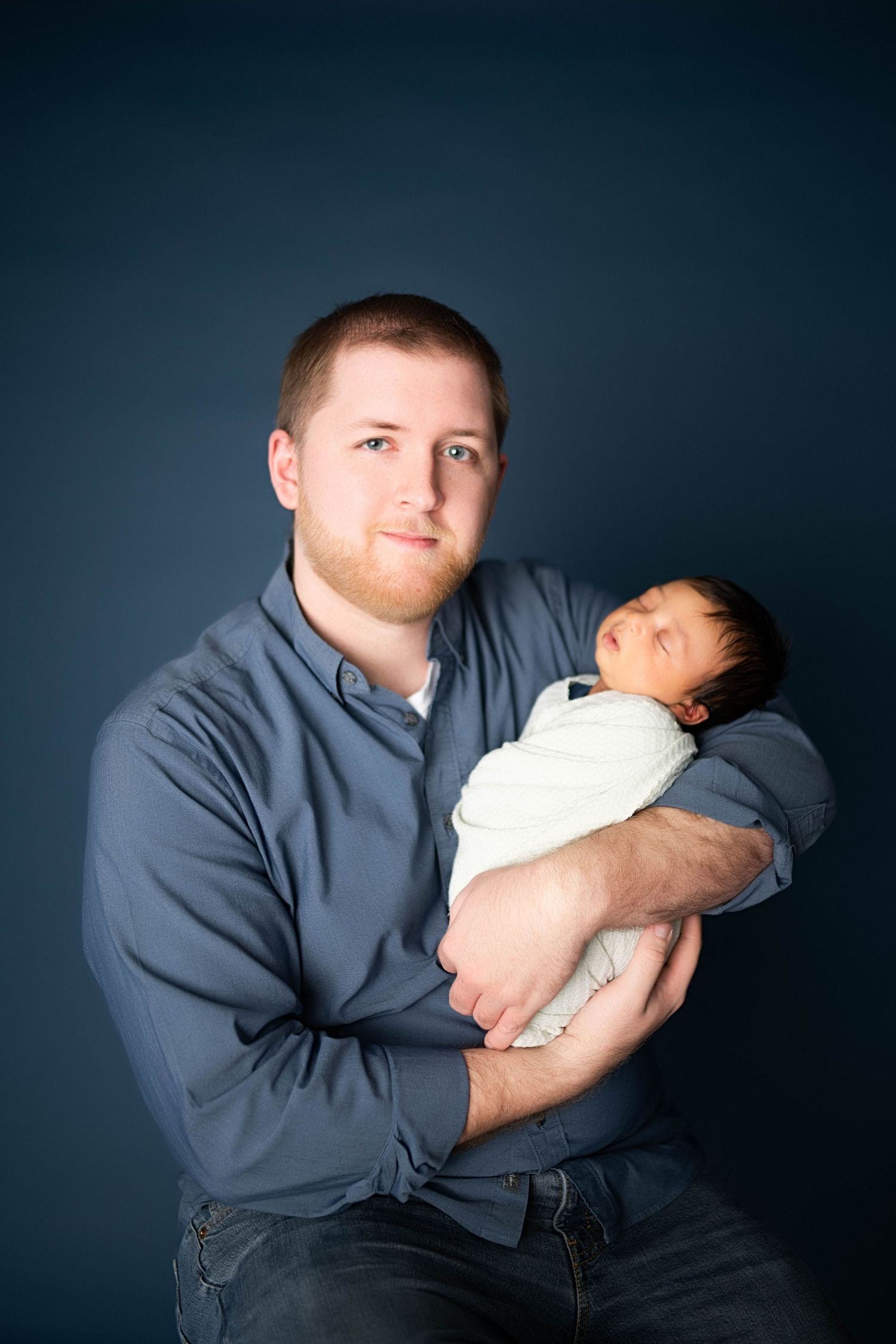 A proud father in a blue shirt holds his swaddled newborn baby against a dark navy background.