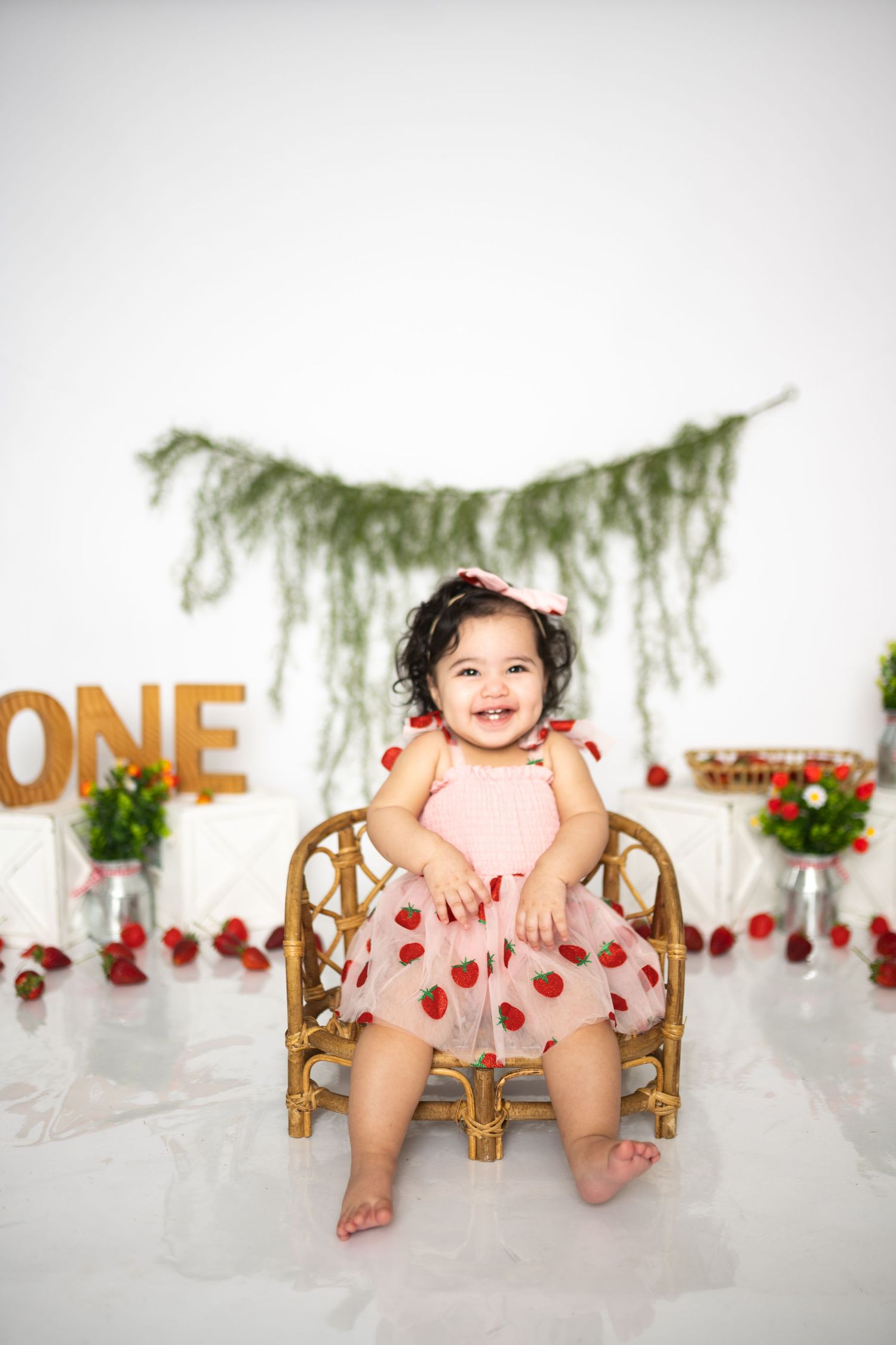 First birthday photoshoot setup with a small wooden chair, pink outfit, and strawberry decorations against white backdrop.