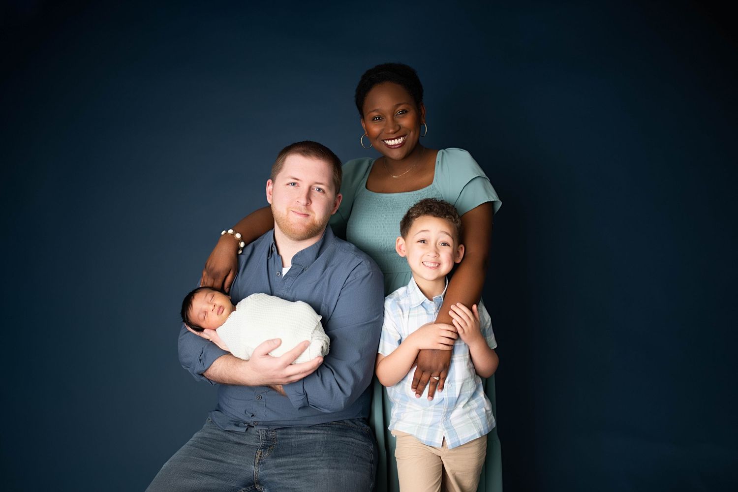 Family portrait with parents holding newborn and toddler son, smiling against a dark blue background.