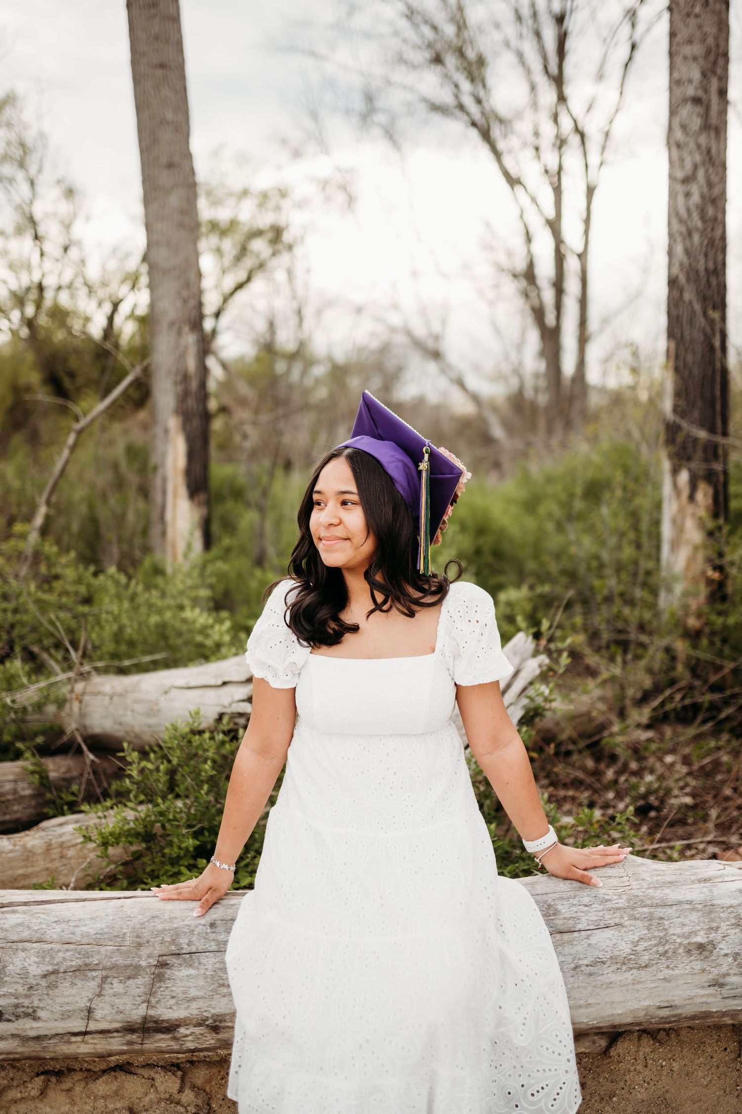 Graduate in white dress sits on fallen log in wooded setting.