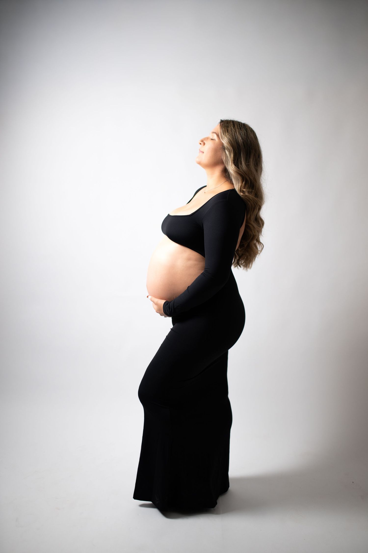 Pregnant woman in black dress cradling her baby bump, posing sideways in a studio maternity photo shoot.