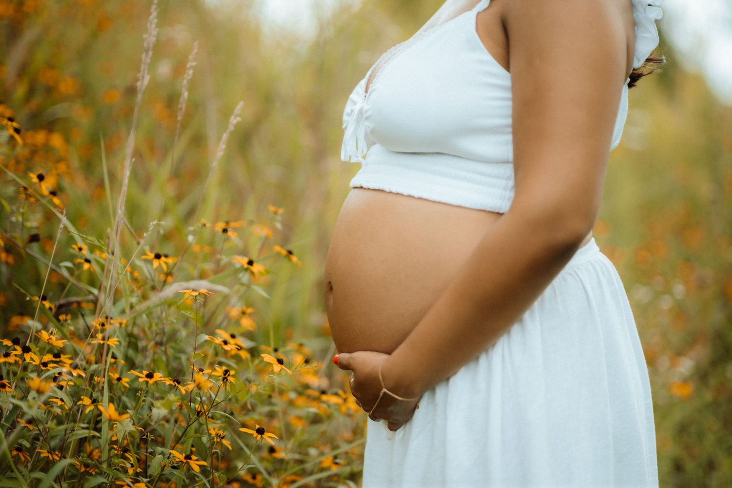 Close-up of pregnant belly in white dress surrounded by golden wildflowers at sunset.