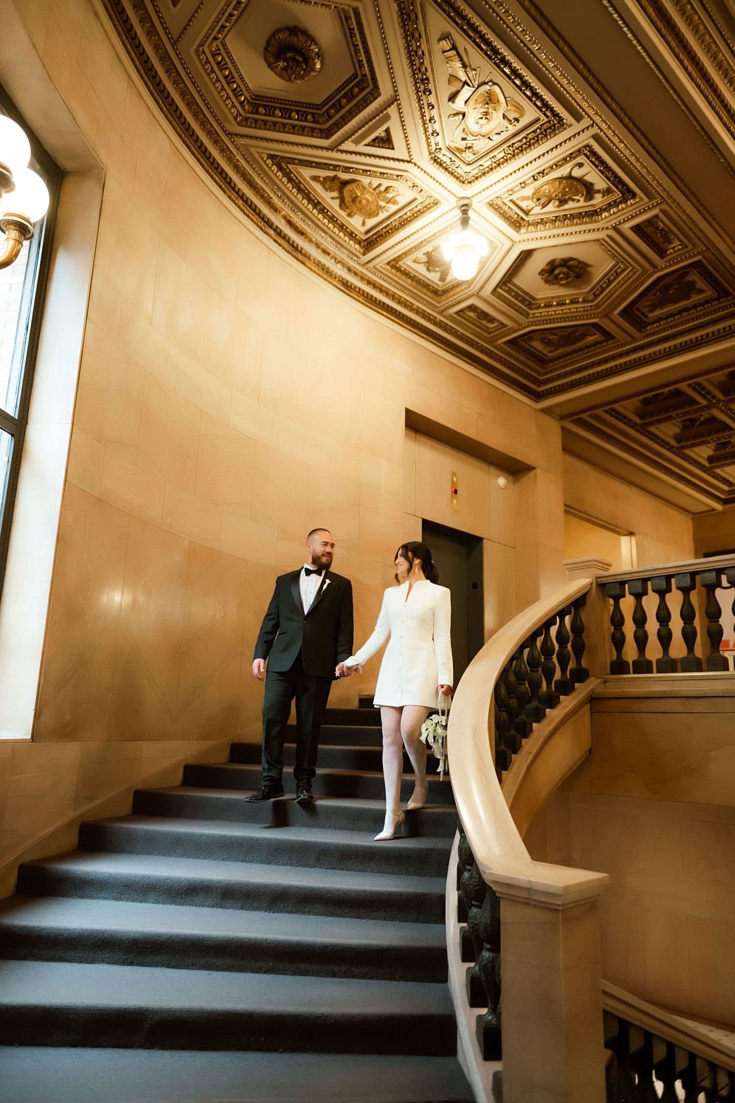 A couple descends an elegant staircase with ornate ceiling details and curved railings in a historic building.