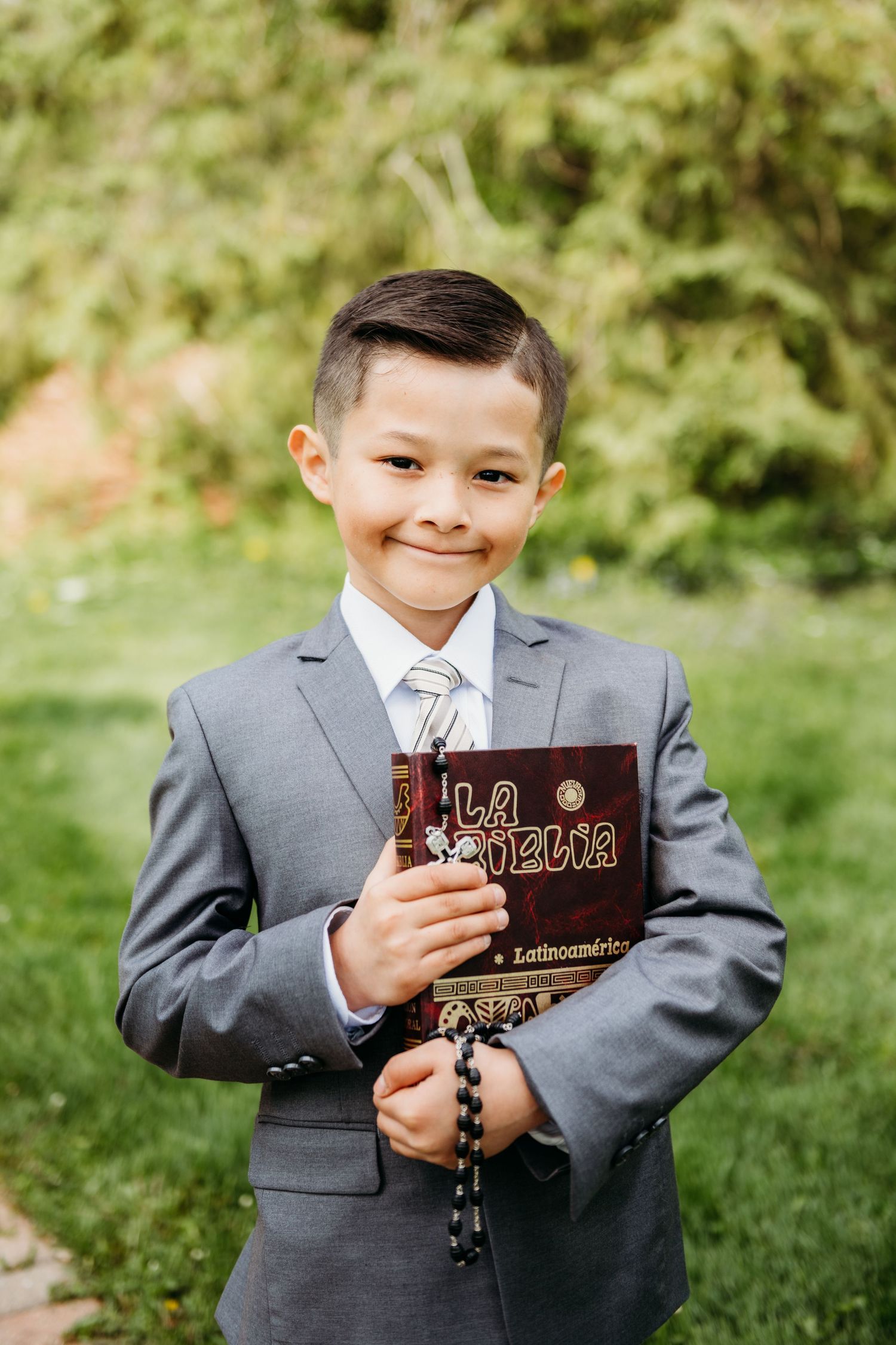 Young boy in formal grey suit smiling while holding religious book outdoors.