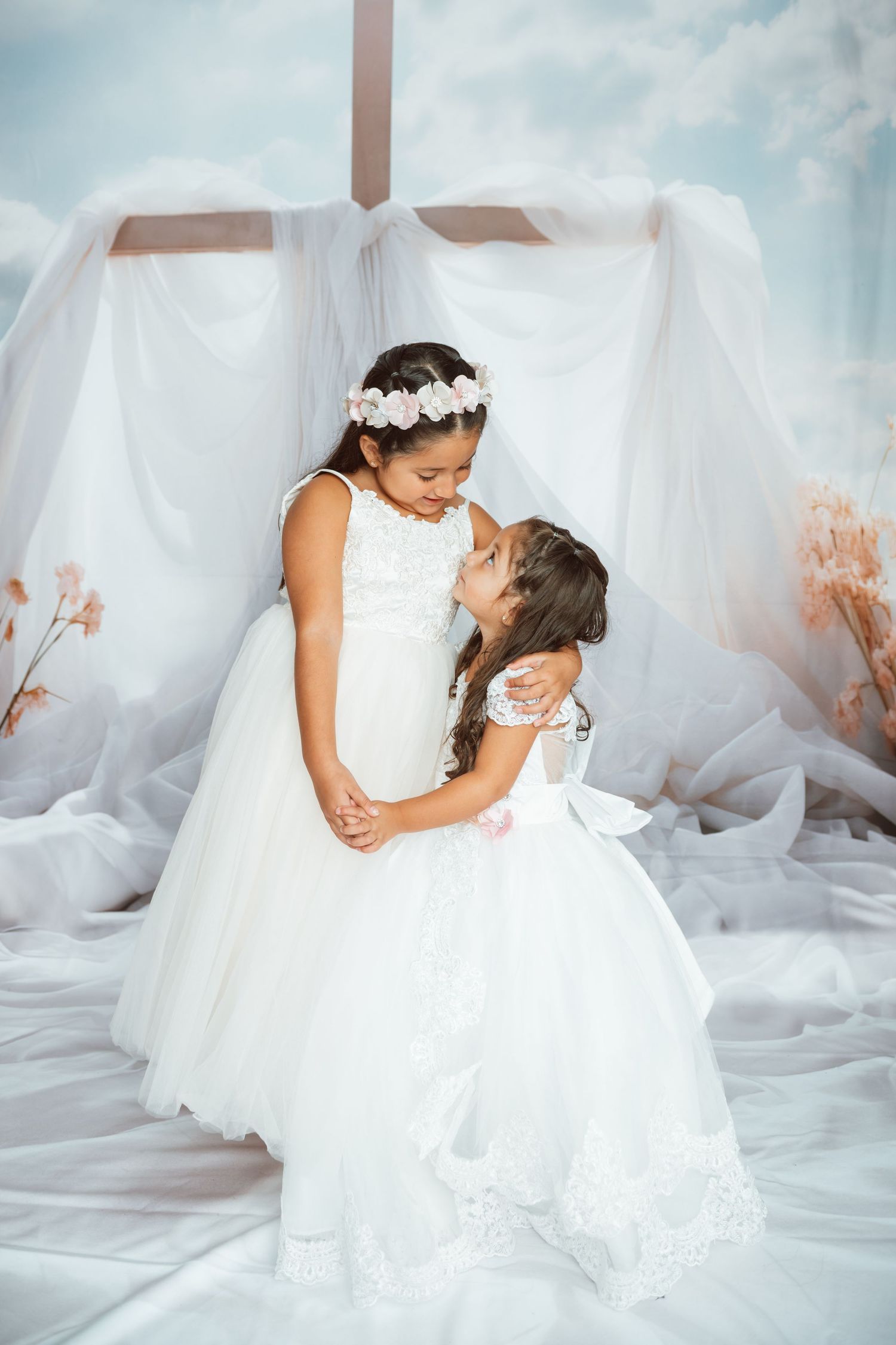 Children in matching white dresses sharing a tender moment against soft blue background.