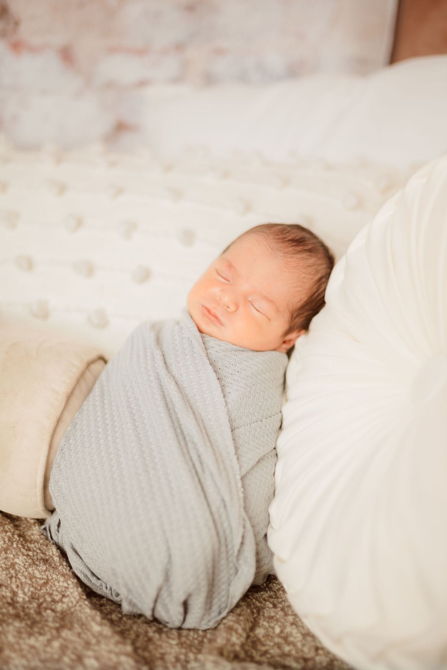 Peaceful newborn baby swaddled in a soft gray wrap, sleeping soundly on a cozy bed with white pillows.