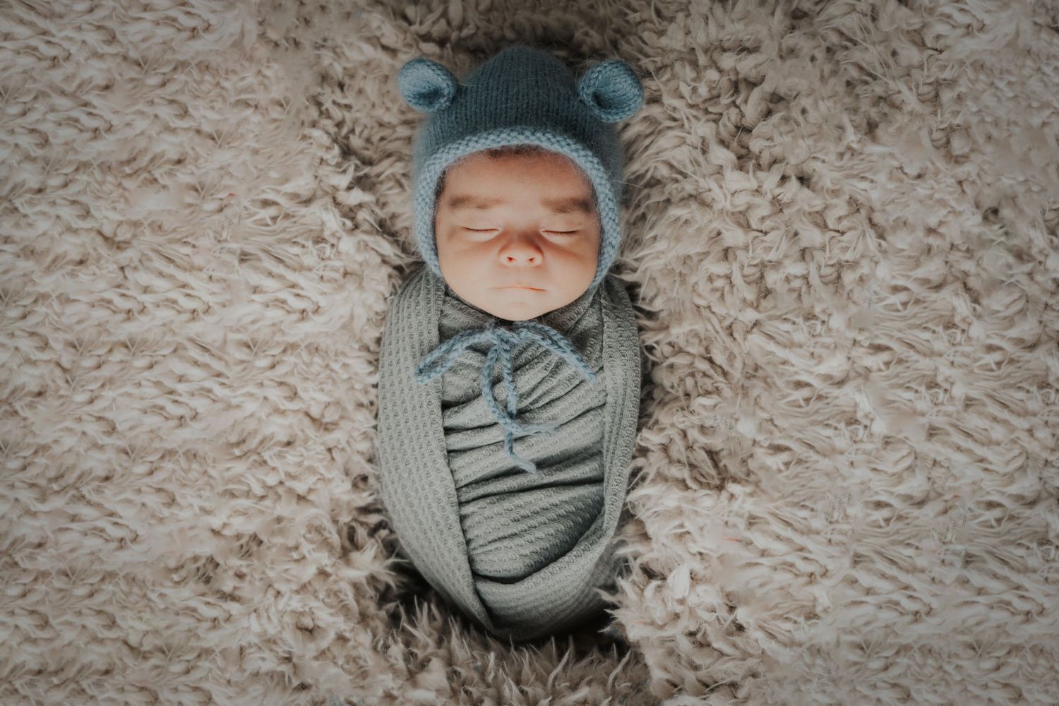 Newborn baby sleeping peacefully swaddled in a blue knit wrap and bear hat on a fluffy beige rug.