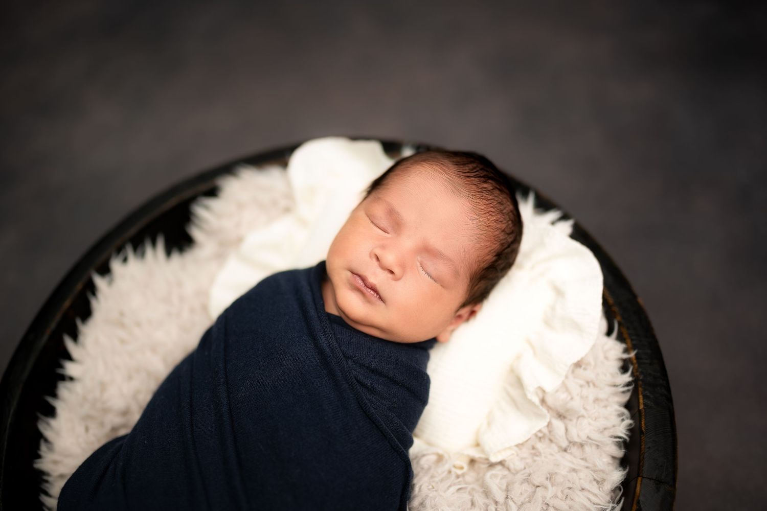 Sleeping newborn baby swaddled in navy blue wrap resting in a round black bowl with white fur lining.