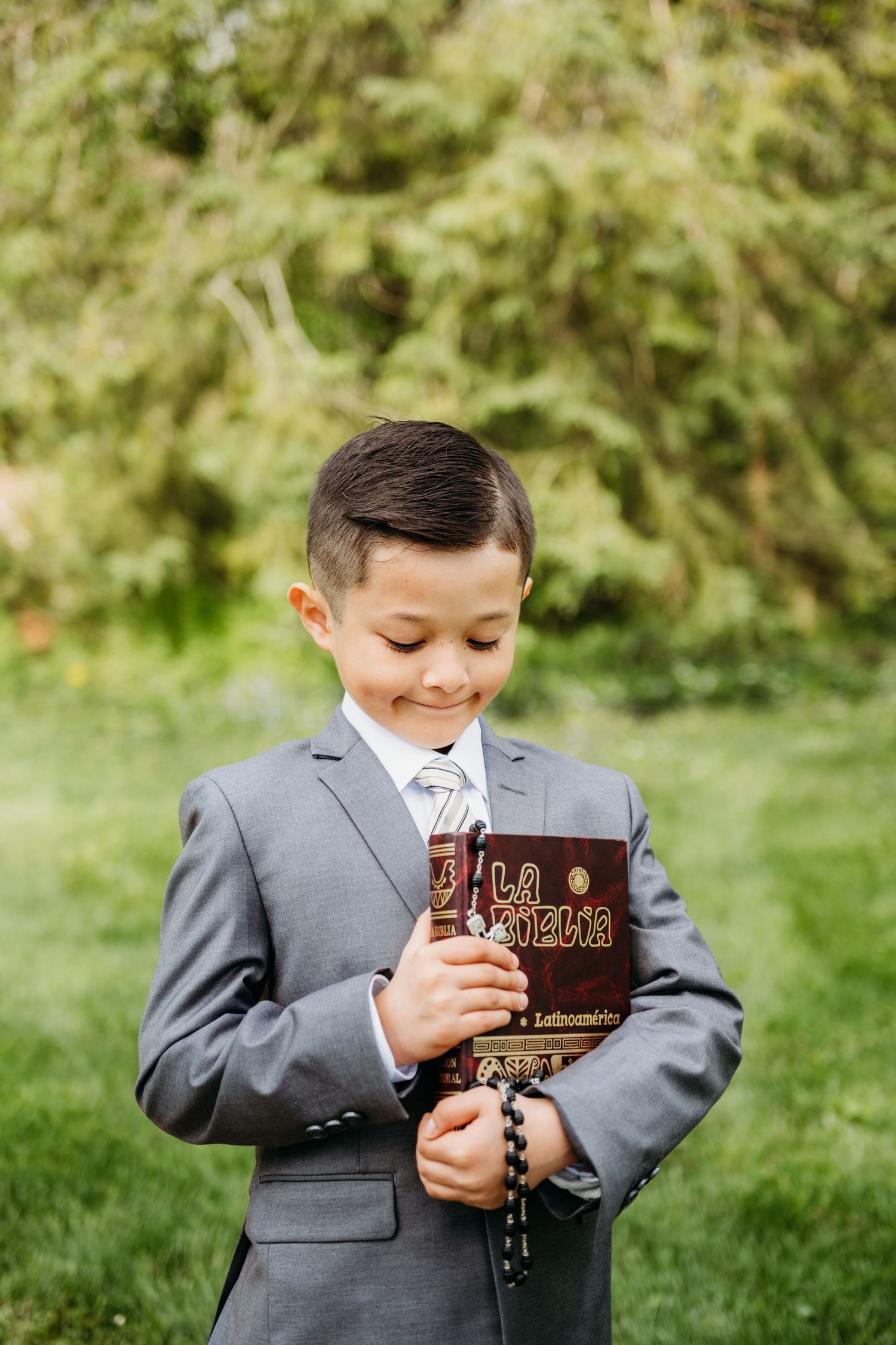 Child in grey suit holding prayer book outdoors in natural setting.