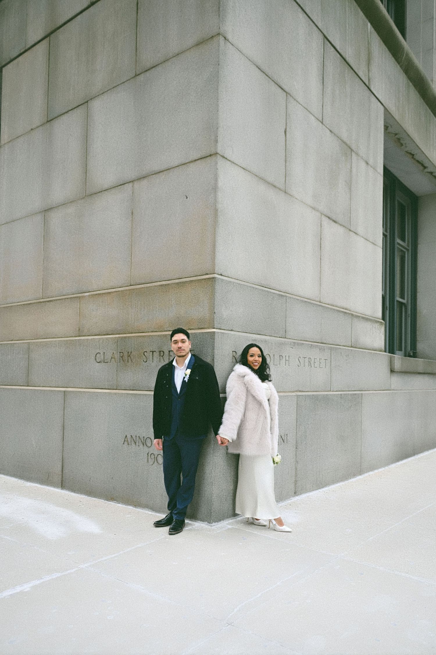 A bride and groom pose together outside City Hall against a stone wall.