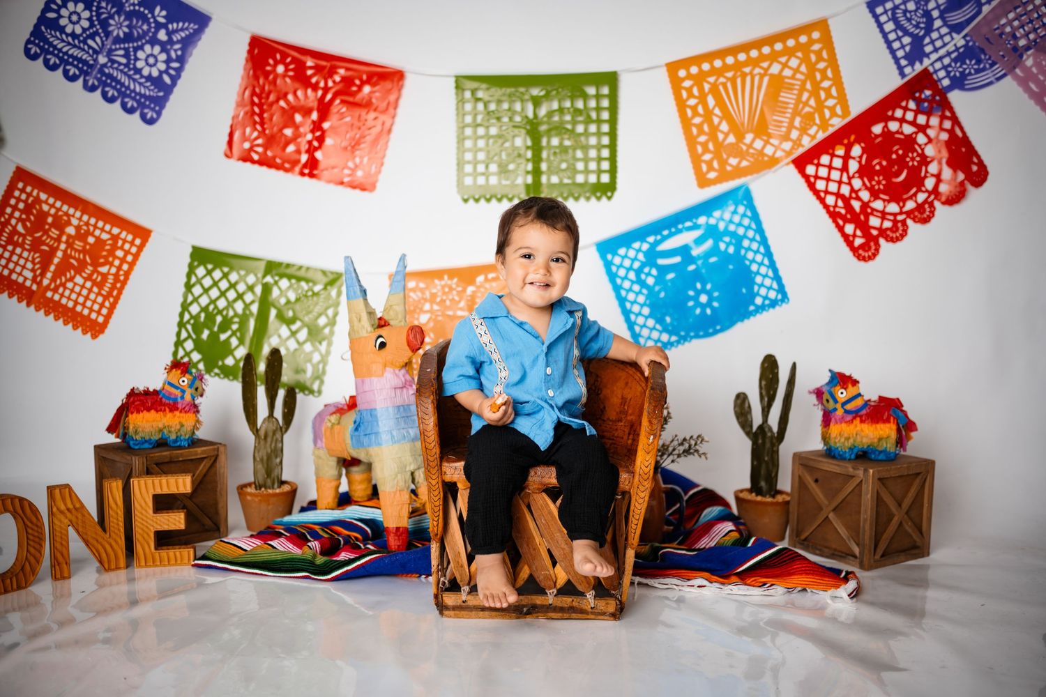 Colorful papel picado banners hang above a festive cake smash setup with wooden decorations.