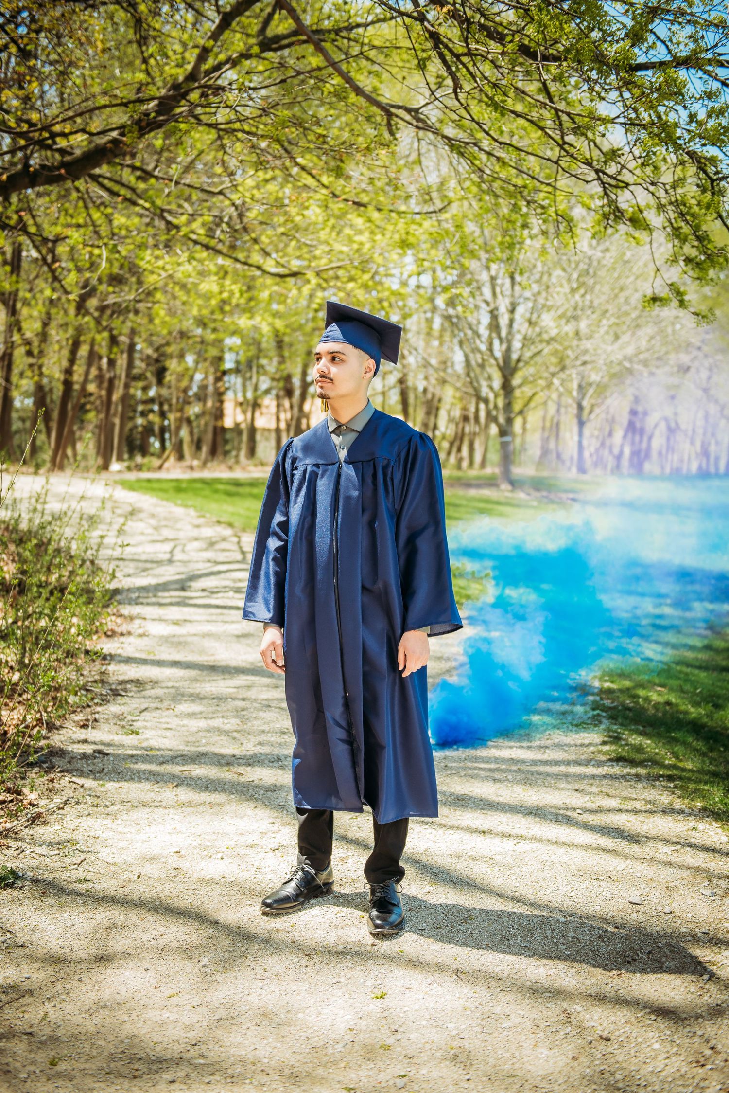A graduate in blue cap and gown stands amid blue smoke on a sunny forest path.
