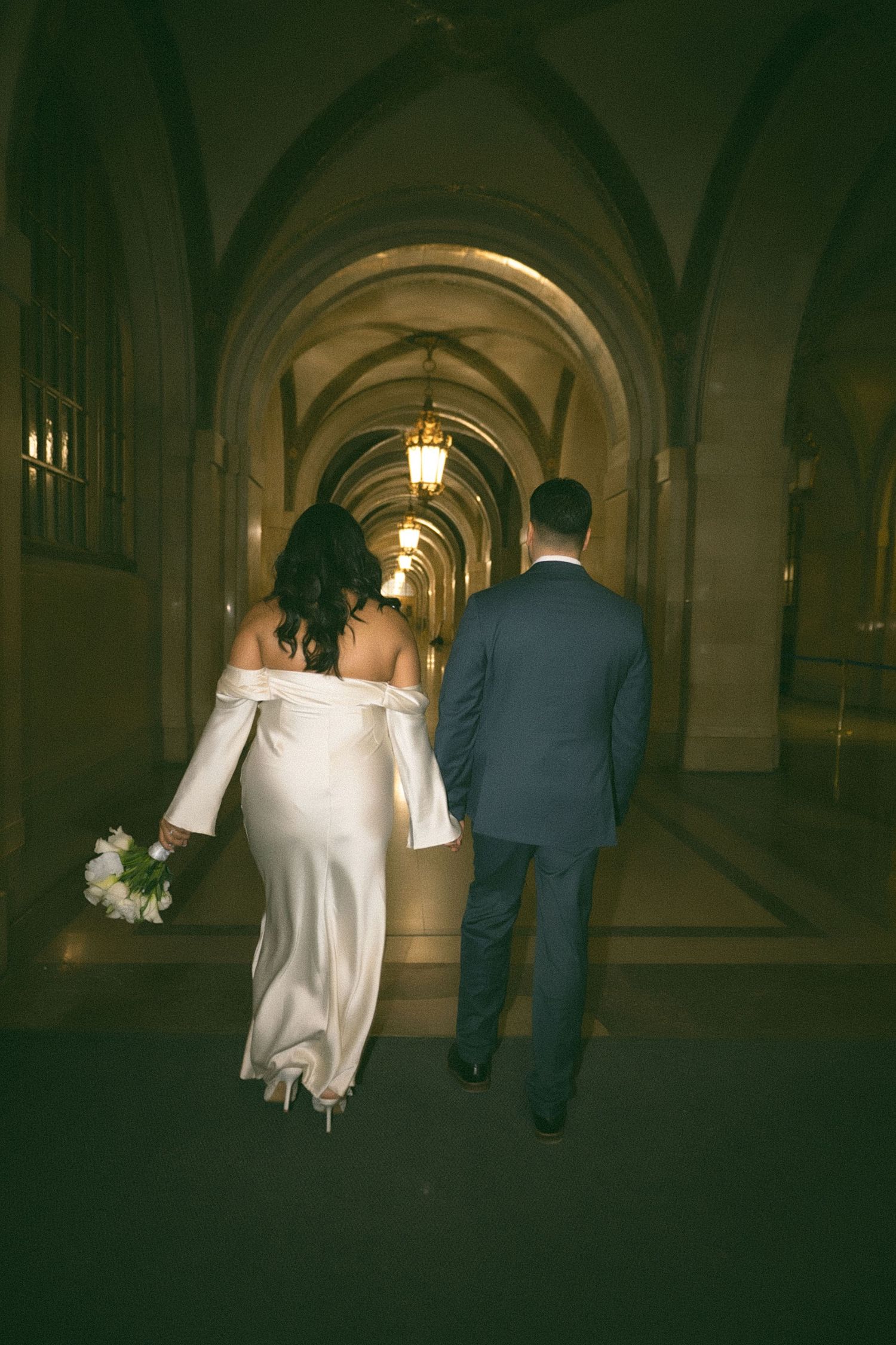 A couple walks through a dimly lit arched hallway in formal attire, creating a romantic silhouette.