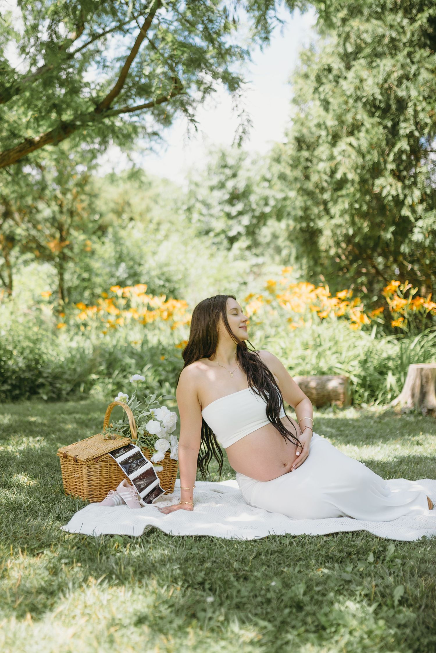 Pregnant woman in white tube top sits on blanket at outdoor picnic with wicker basket and ultrasound photos.