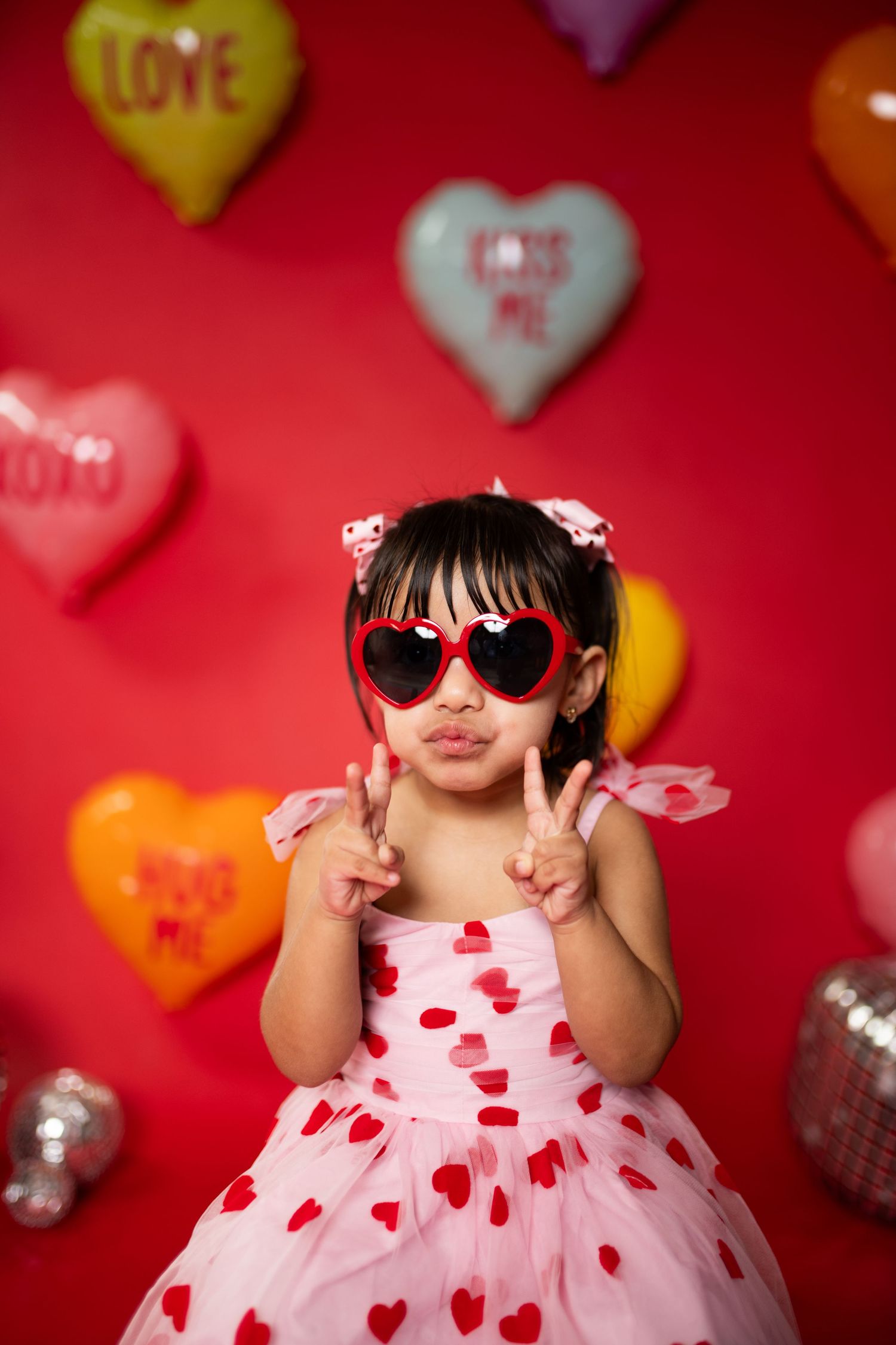 Little girl in heart-patterned pink dress and sunglasses poses against red Valentine's backdrop.