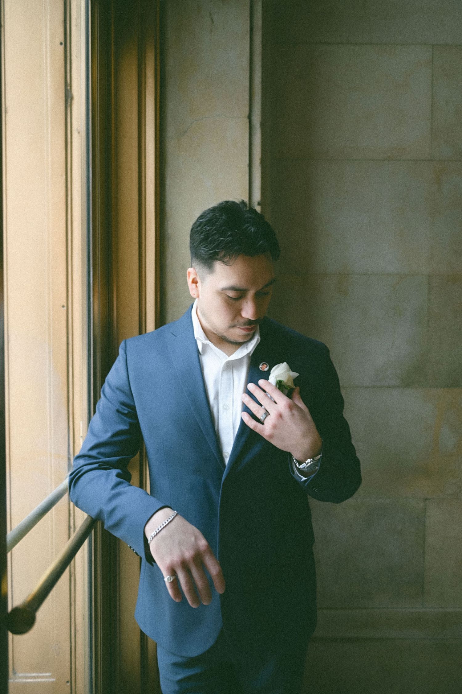 A groom in a navy suit adjusts his tie while standing near wooden doors.