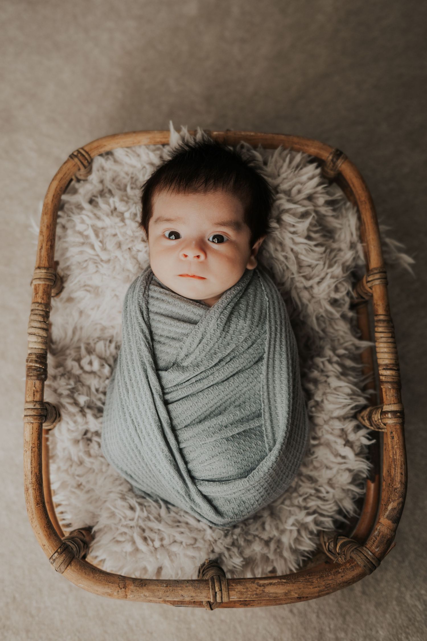 Newborn baby swaddled in sage green wrap lying in a rustic wicker basket with fluffy fur lining.